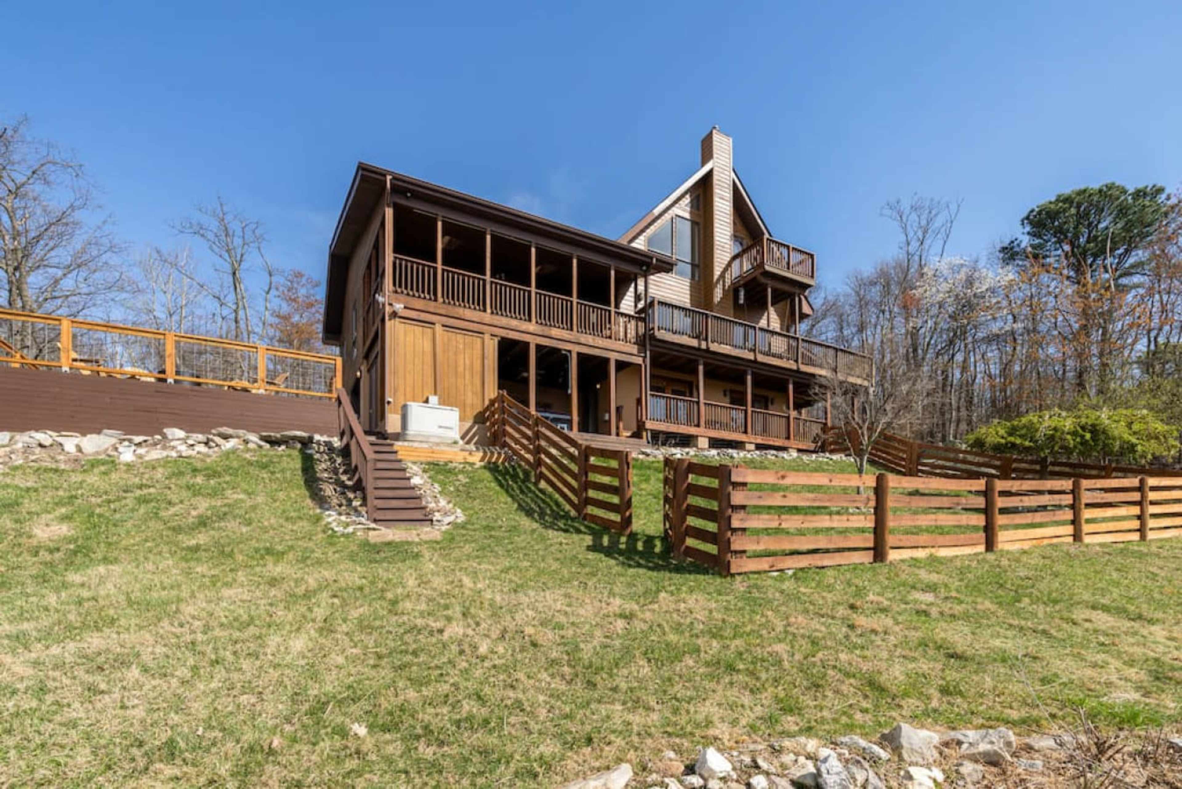 A two-story wooden house located on a sloped lawn, with a fenced yard and a deck overlooking the surroundings.