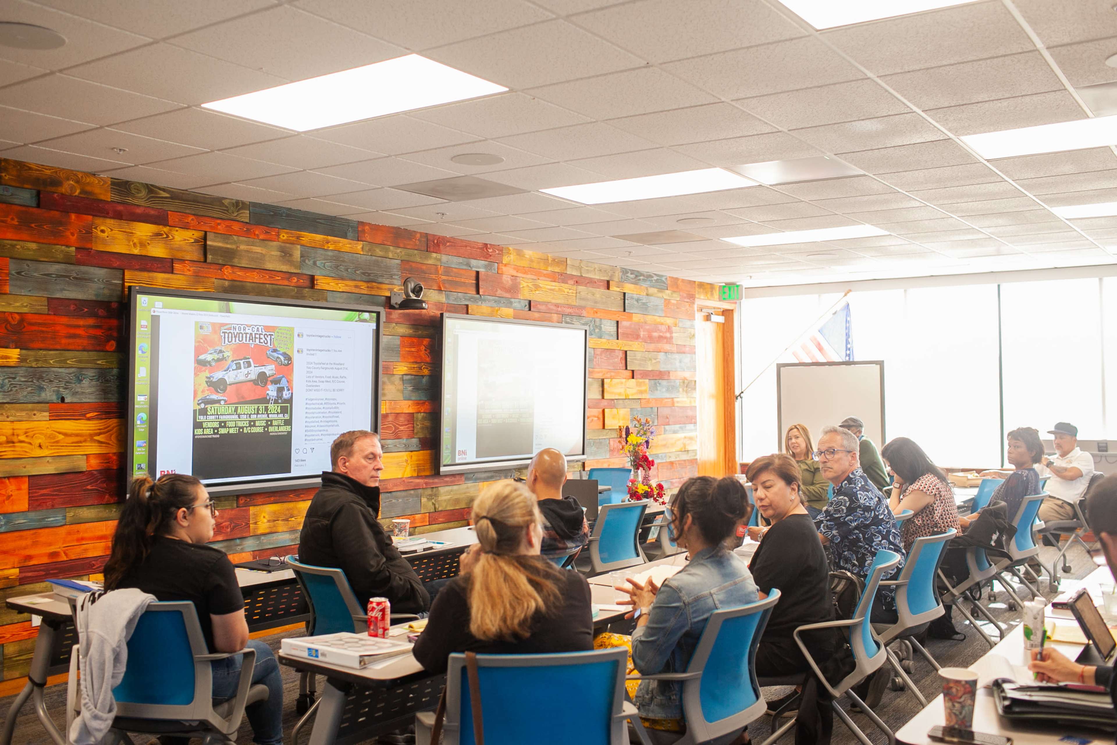 A group of people is seated in a classroom with wooden-paneled walls, engaging in discussion while facing a screen displaying promotional material.
