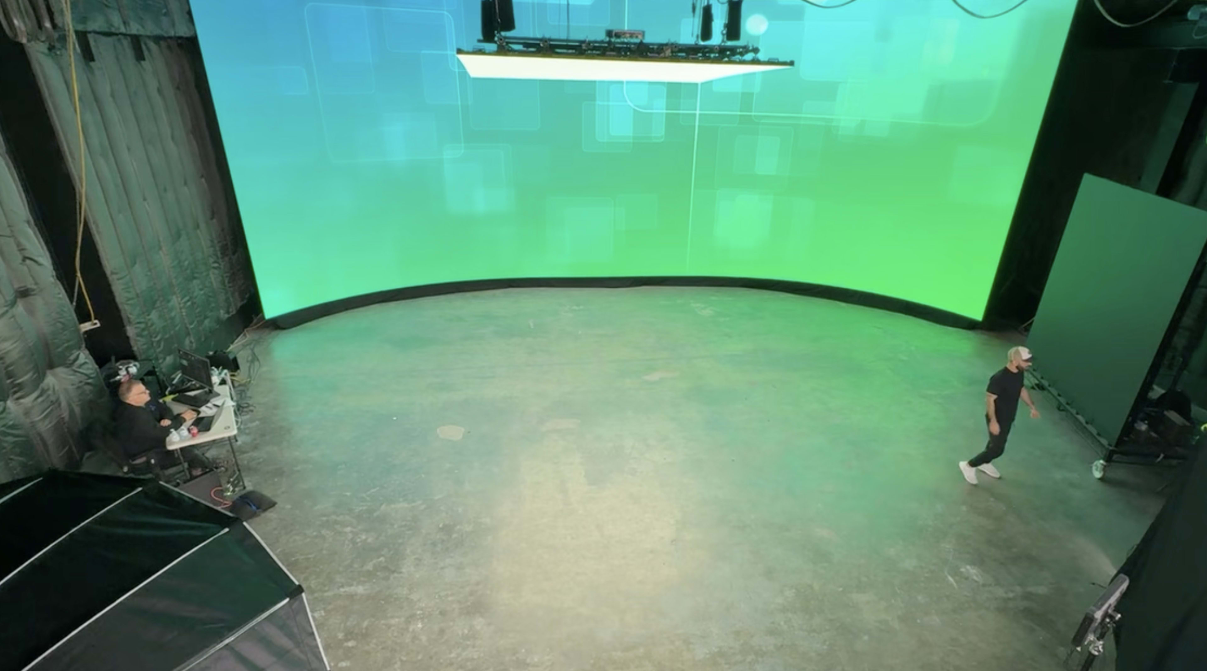 A person stands on a large, empty stage with a curved green screen backdrop, while another person works at a desk with equipment on the side.