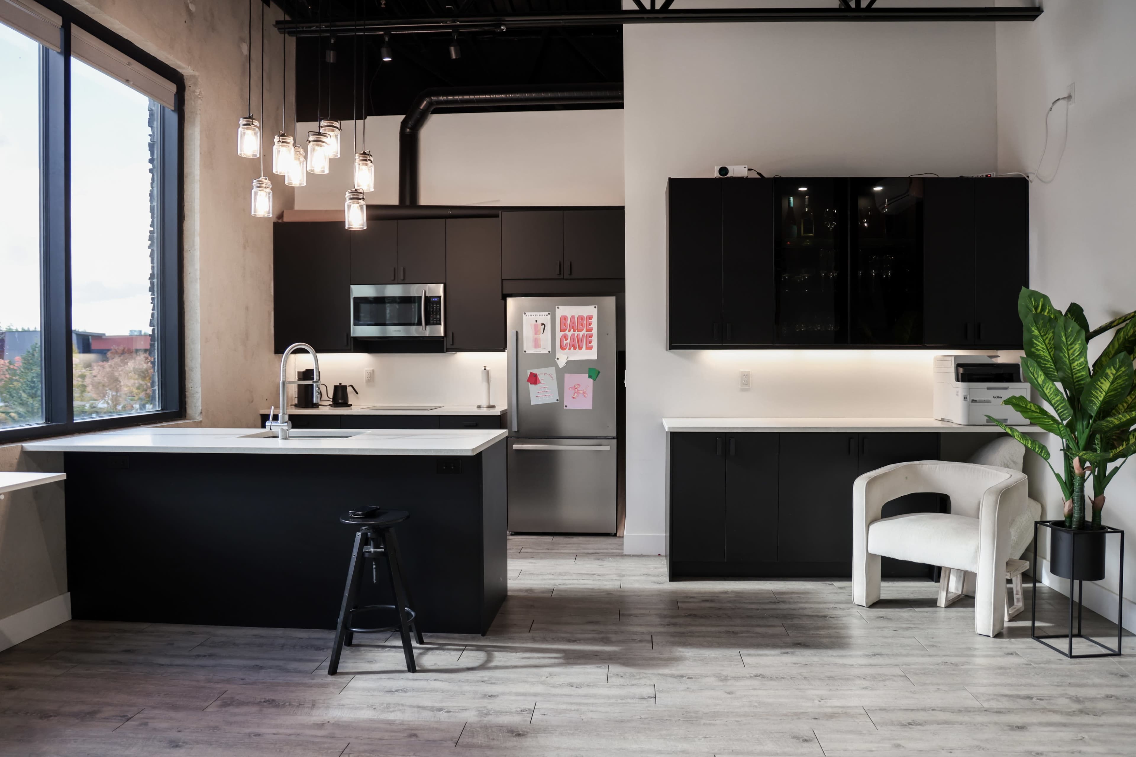 The image shows a modern kitchen with black cabinets, a central island, and large windows allowing natural light to enter the space.
