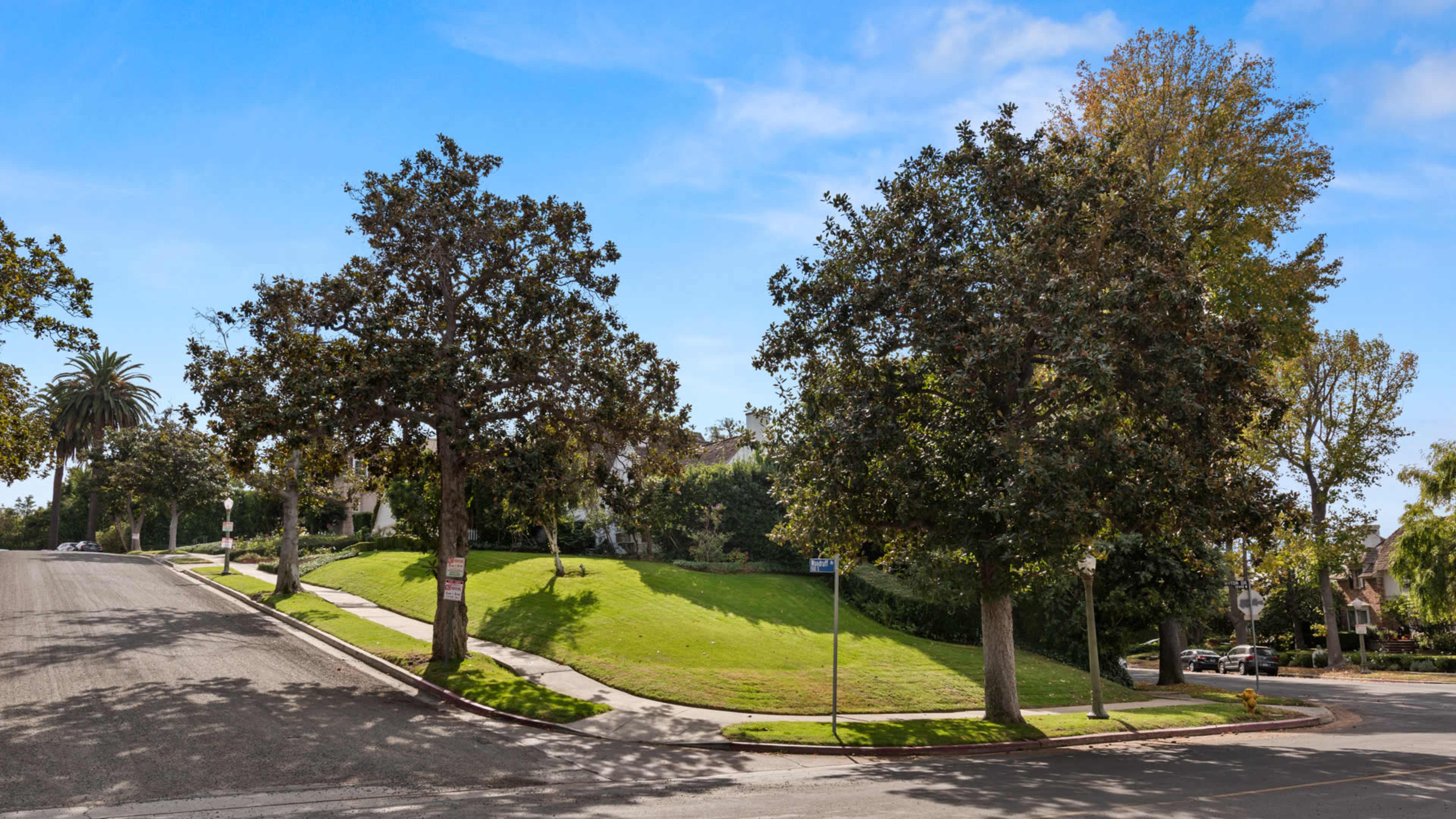A grassy, sloped corner lot bordered by several trees and a quiet street.