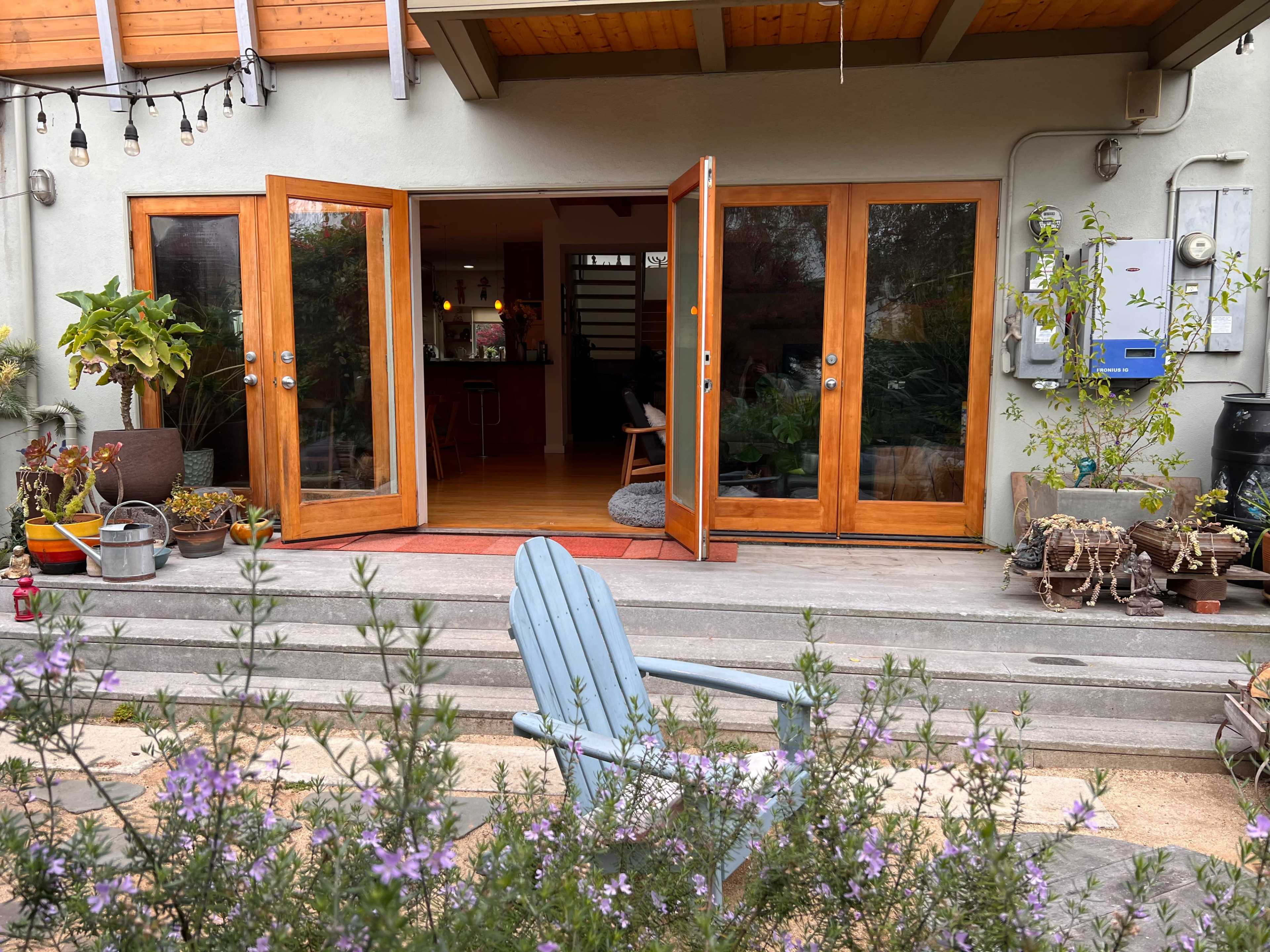 A patio area with opened double doors leading into a home, a blue wooden chair, and various potted plants and decor lining the entrance.