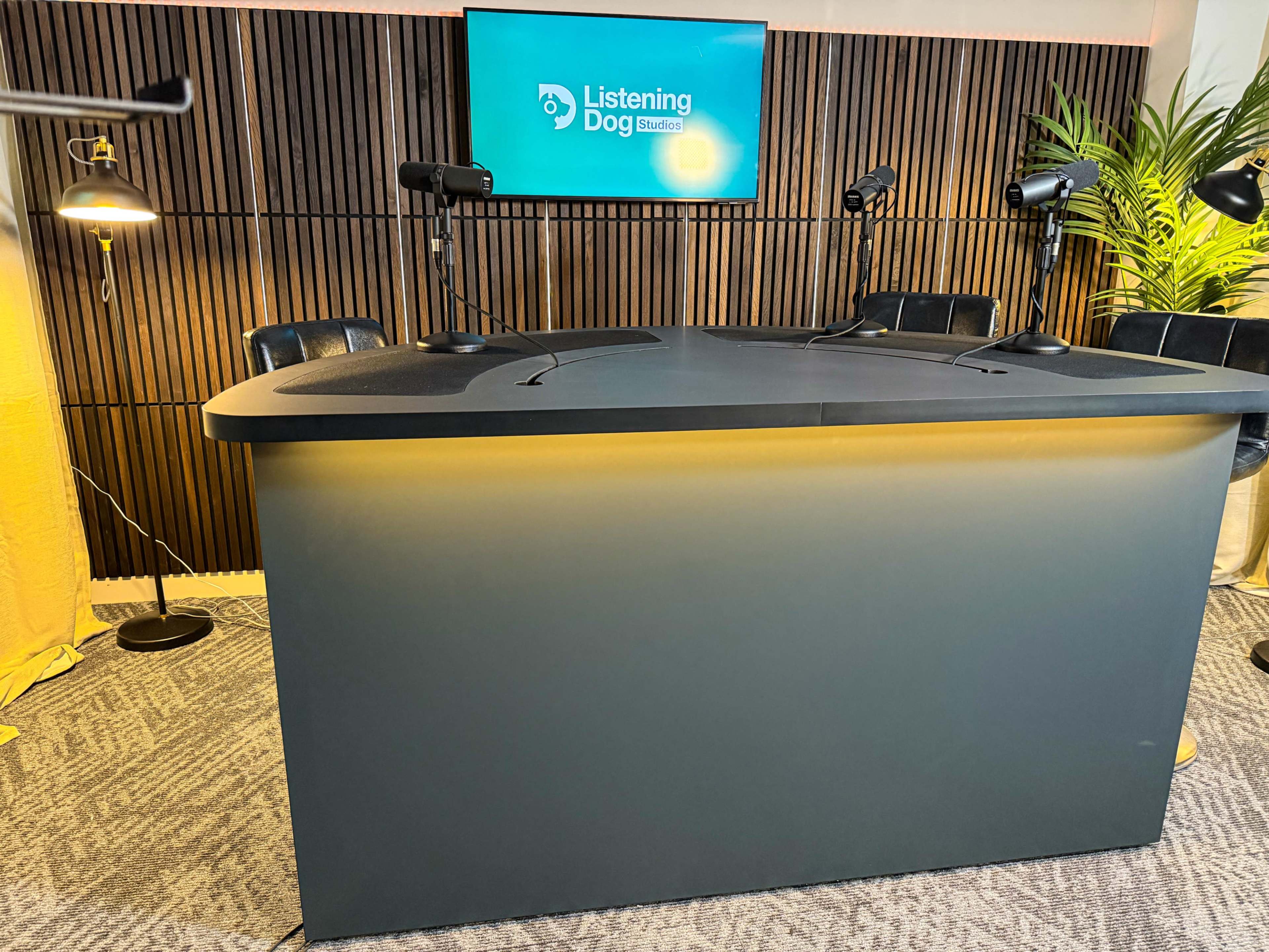 The image shows a podcast studio setup with a modern table, microphones, and a screen displaying the logo "Listening Dog Studios" against a backdrop of wooden paneling and plants.