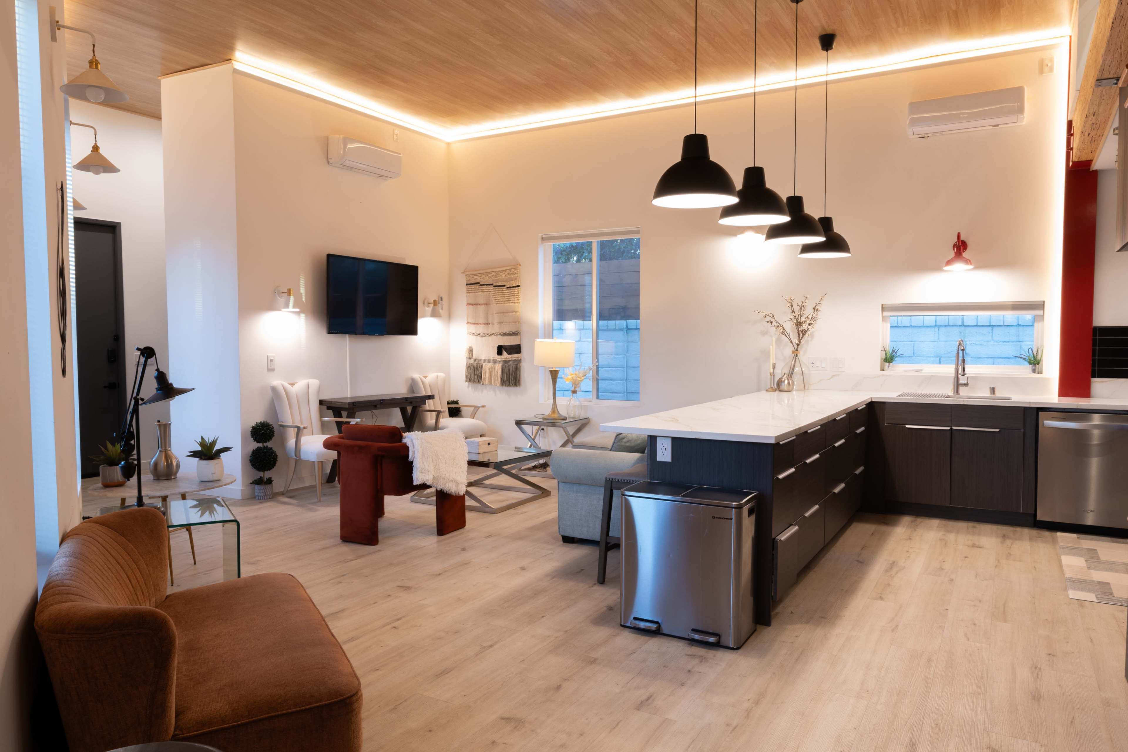 The image shows a modern kitchen and living area featuring a central island with a sink, several pendant lights, and a mix of furniture including a sofa and chairs.