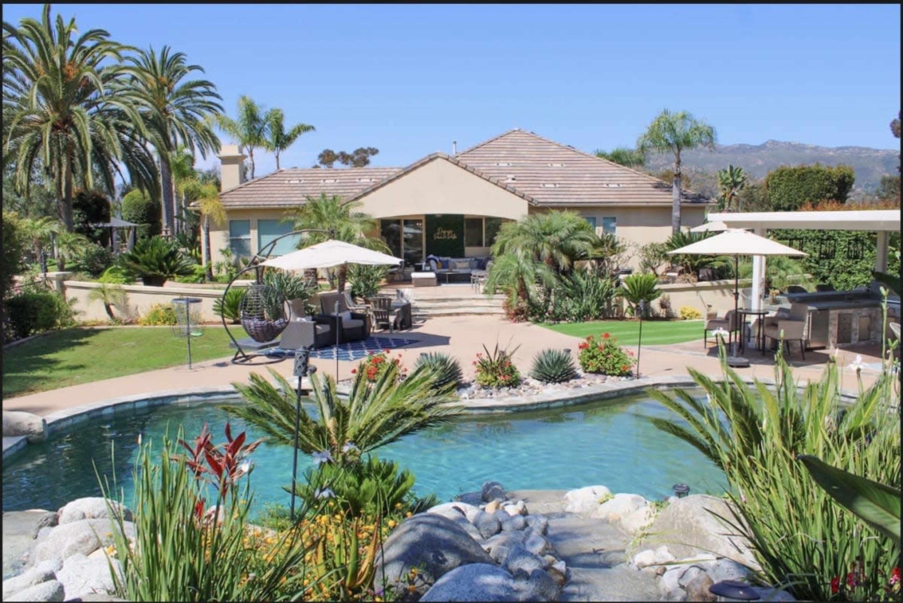 The image shows a landscaped backyard featuring a swimming pool, palm trees, and a house in the background.