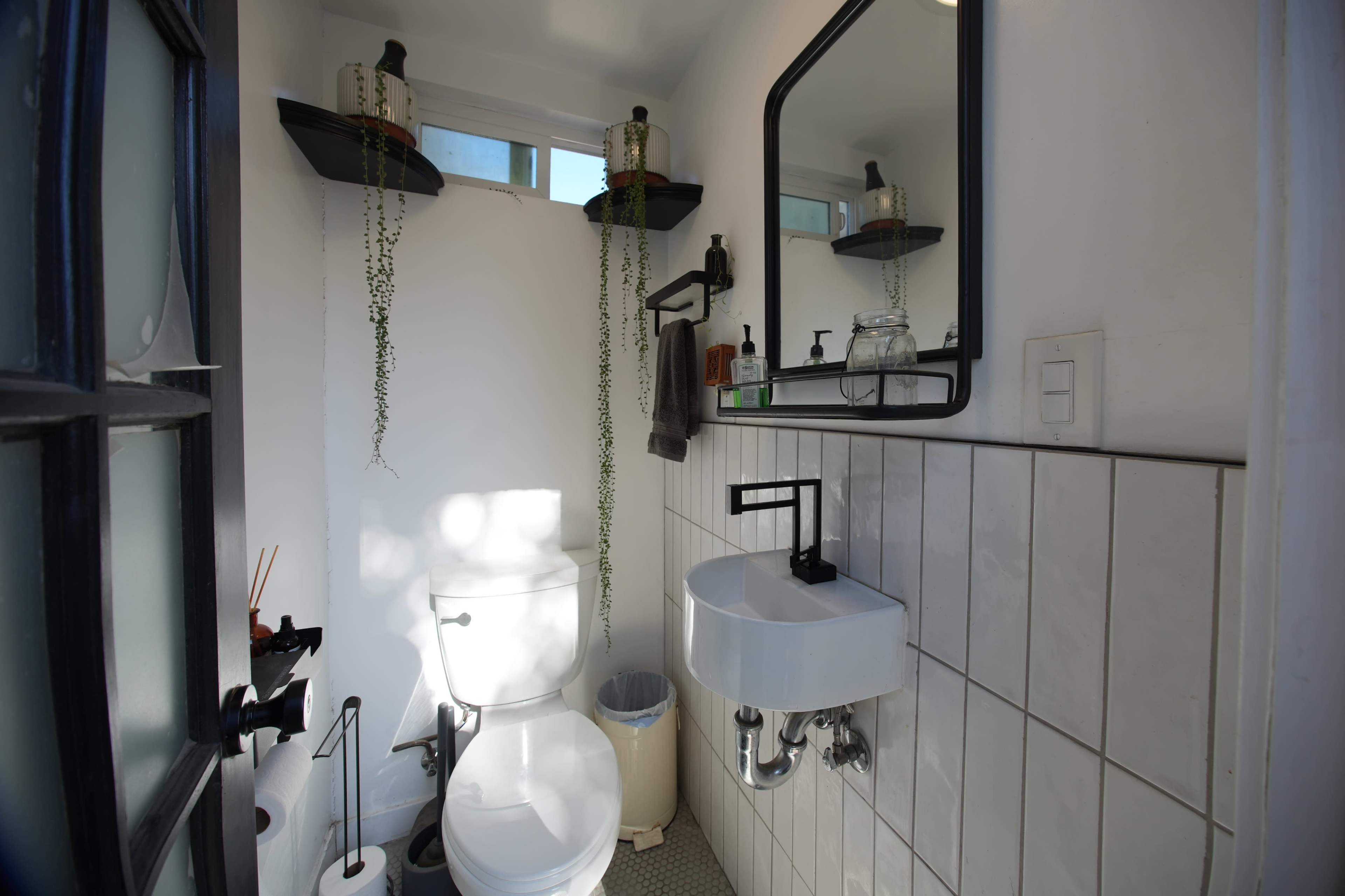 The image shows a small bathroom featuring a white toilet, a wall-mounted sink, and shelves with plants, all set against white walls and tiled flooring.