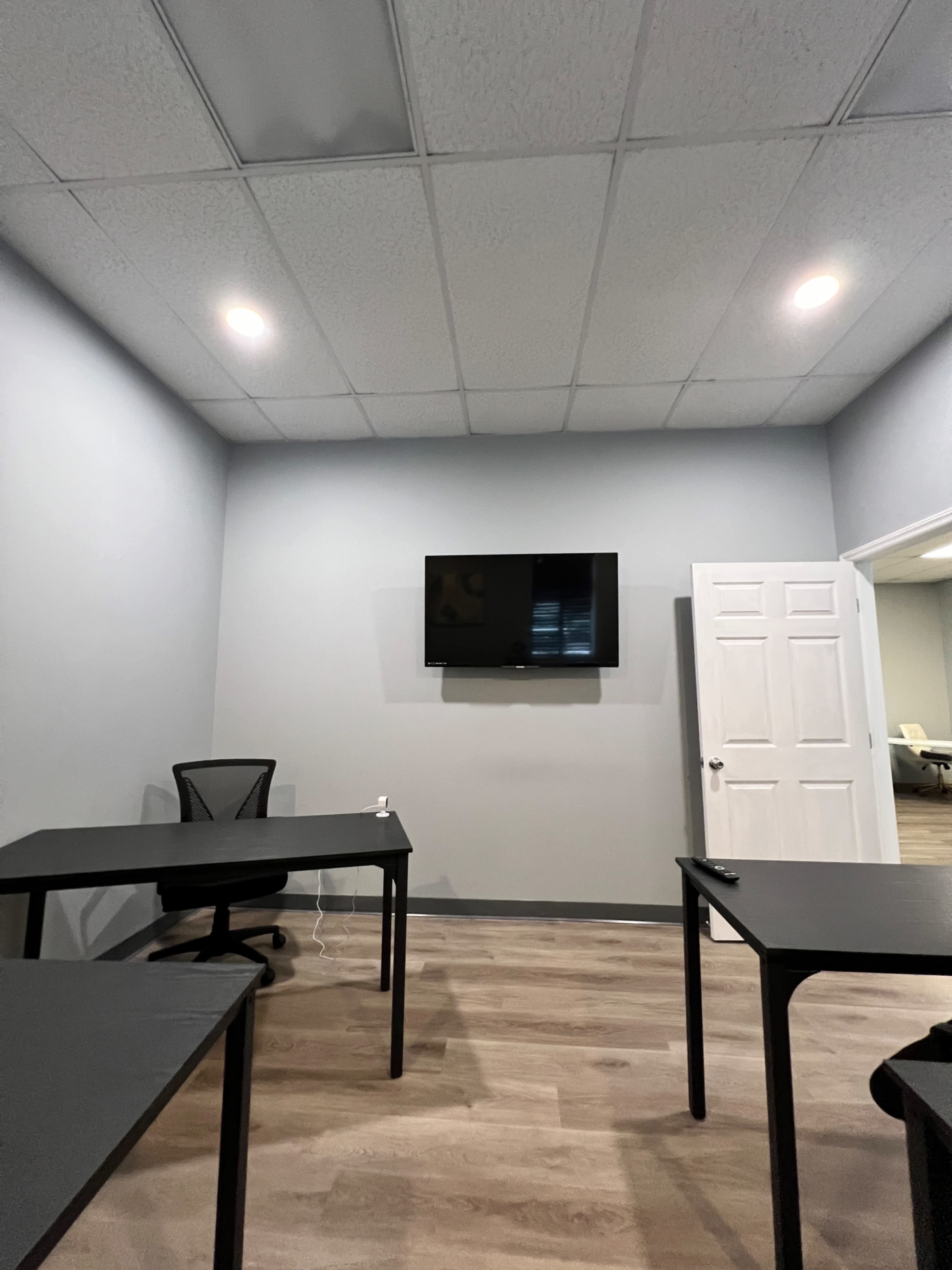 A small meeting room features two black tables, a black chair, and a wall-mounted television, with a door leading to another room in the background.