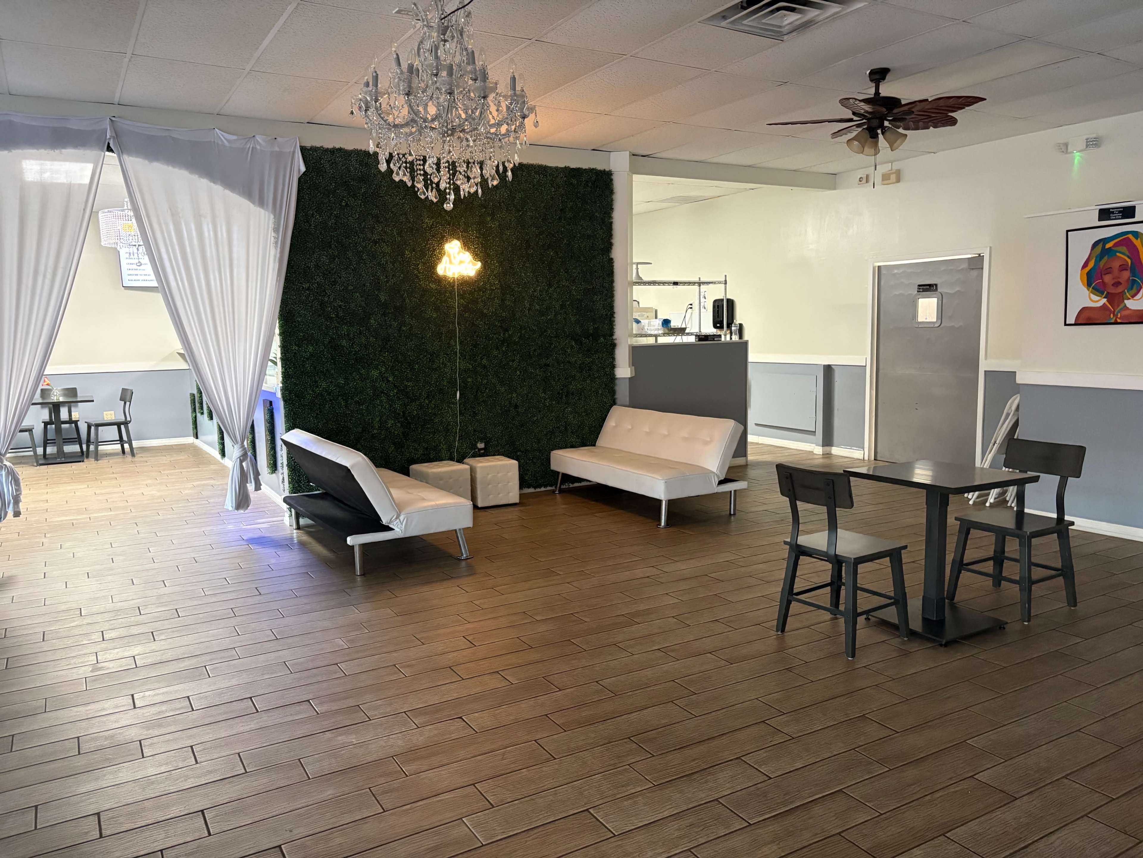 The image shows a spacious indoor area with a mix of modern furniture, including white sofas and a black table with chairs, against a backdrop of a green wall and decorated with a chandelier.