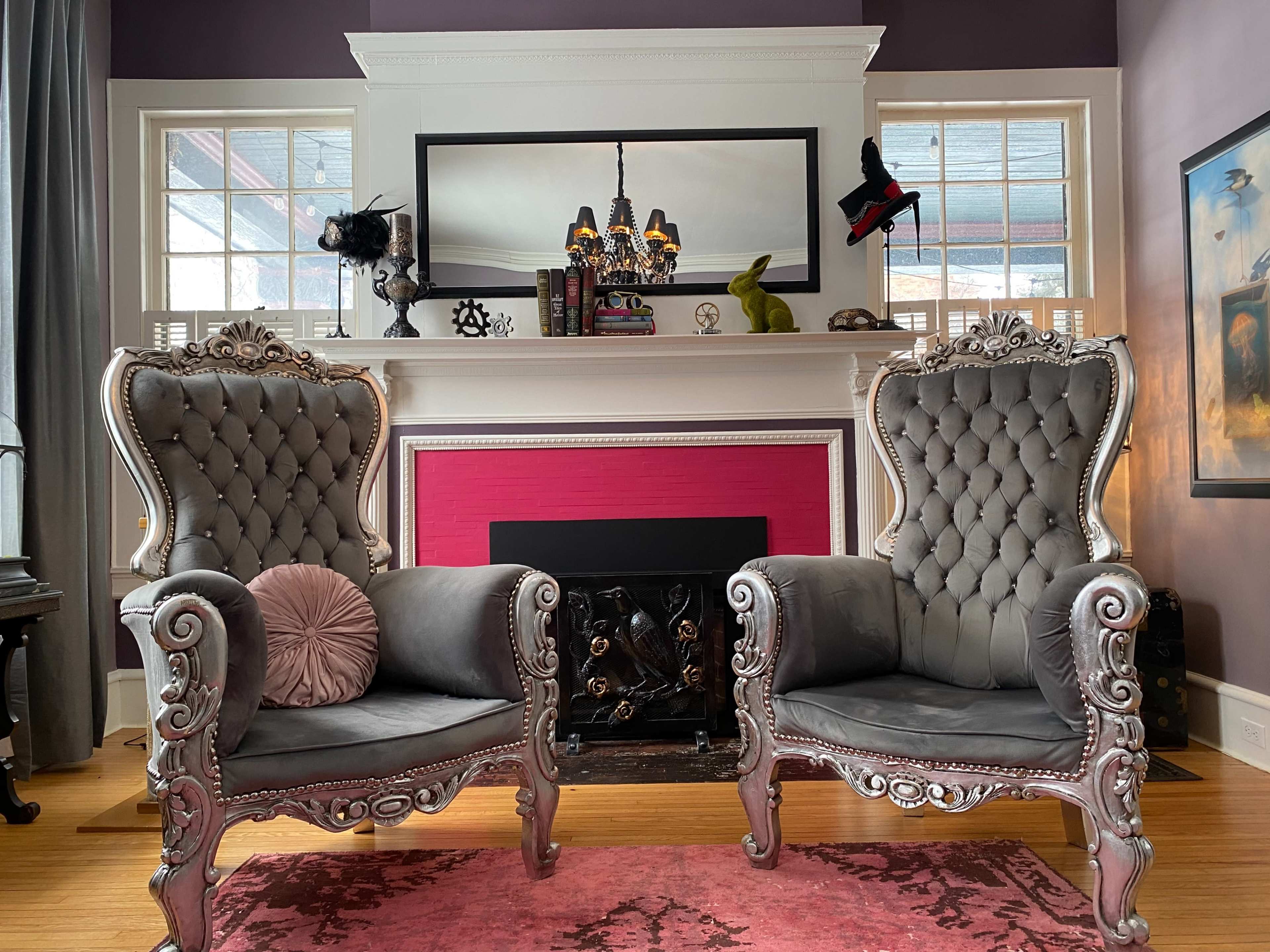 The image shows two ornate, tufted armchairs facing a decorated mantel with a mirror and various decorative items, set against a purple wall.