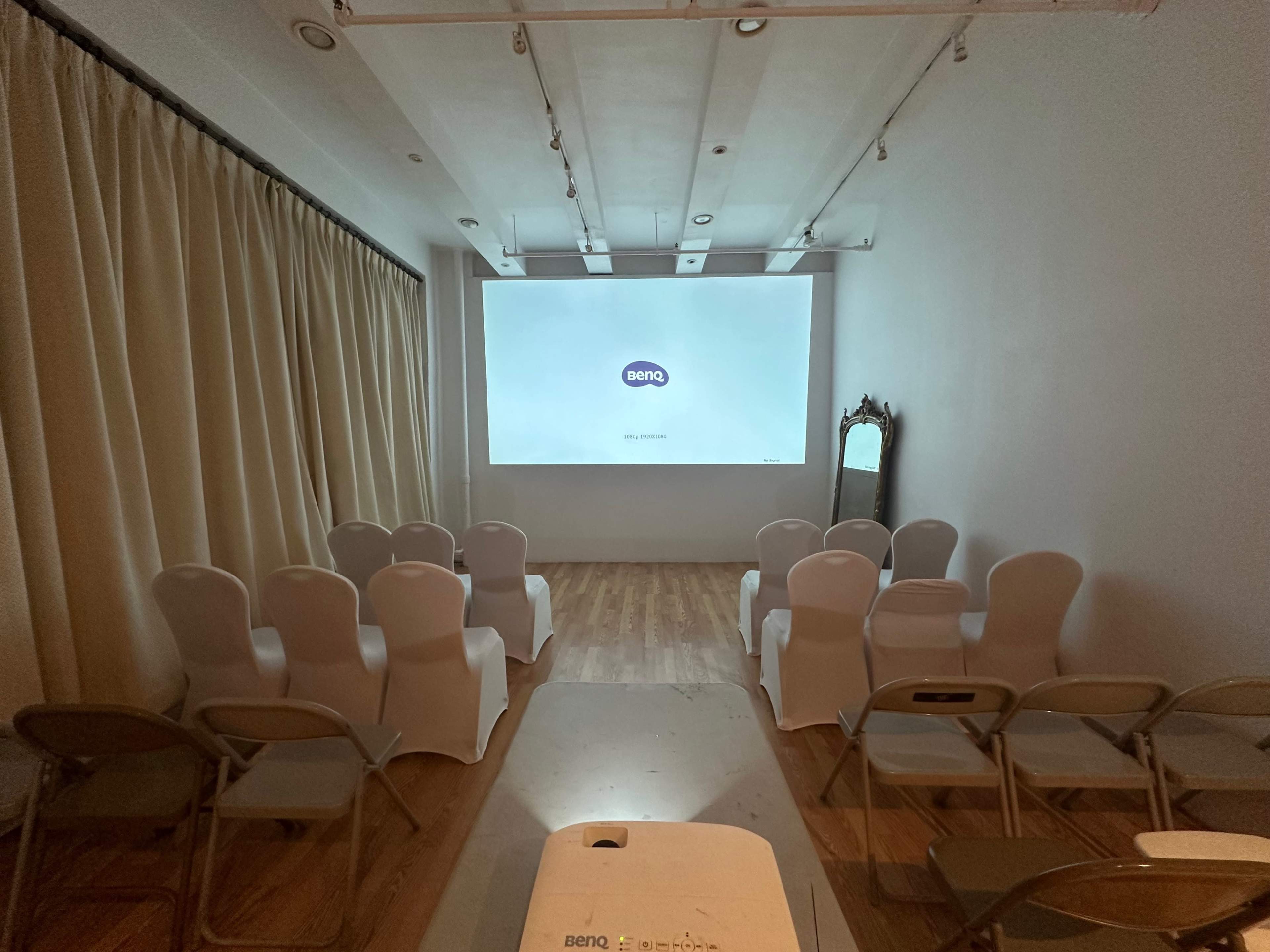 The image shows a minimalist room arranged with rows of chairs facing a projection screen at the front.