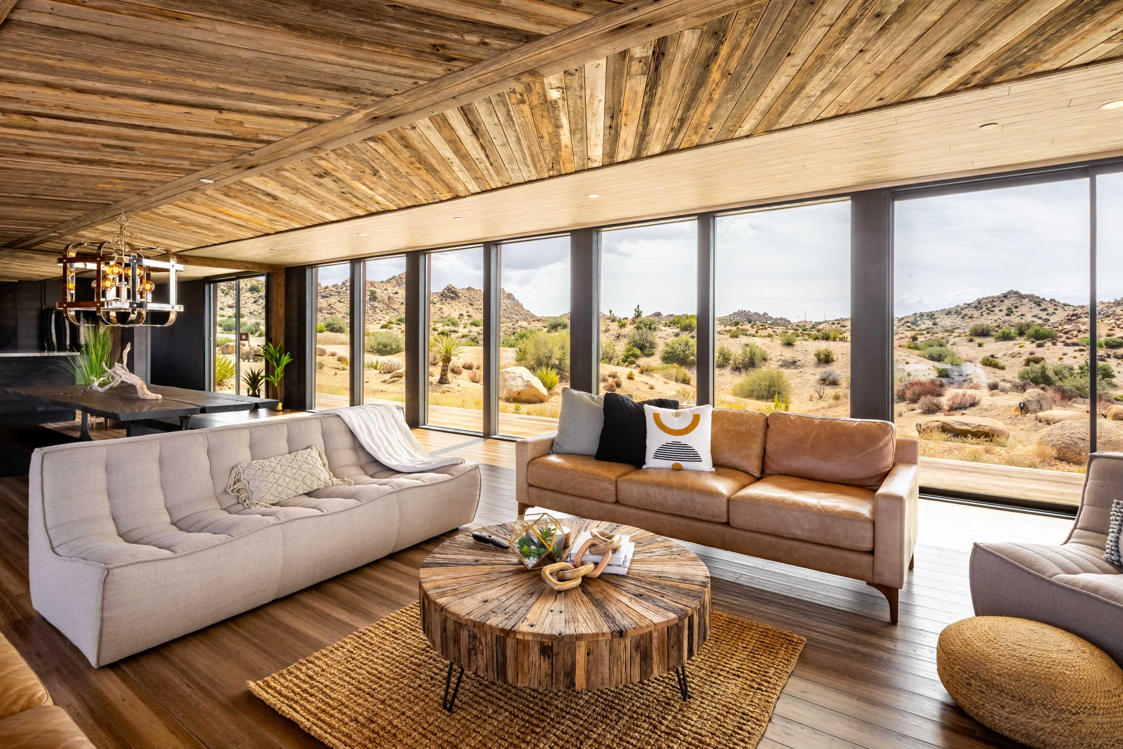 The image shows a modern living room with large windows offering a view of a desert landscape, featuring a mix of light-toned furniture and a circular wooden coffee table.