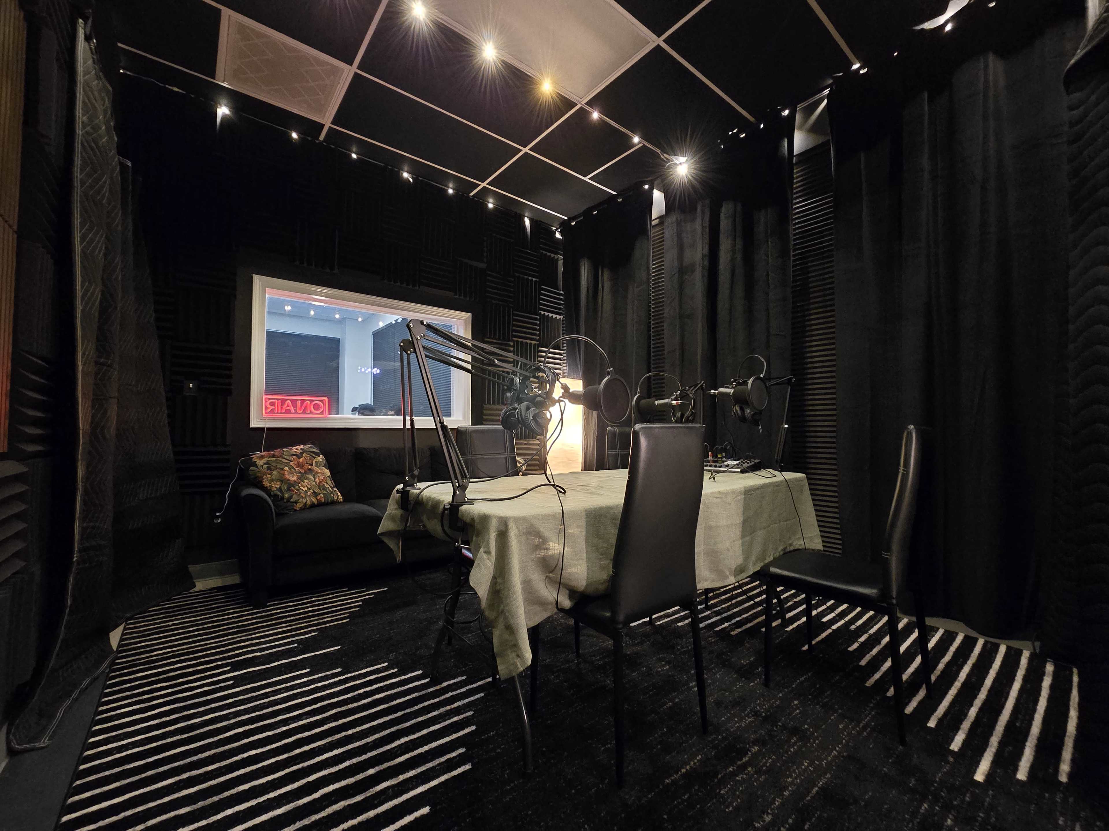 The image shows a soundproof recording studio with a table, chairs, and microphones set up for audio production, surrounded by black acoustic panels.