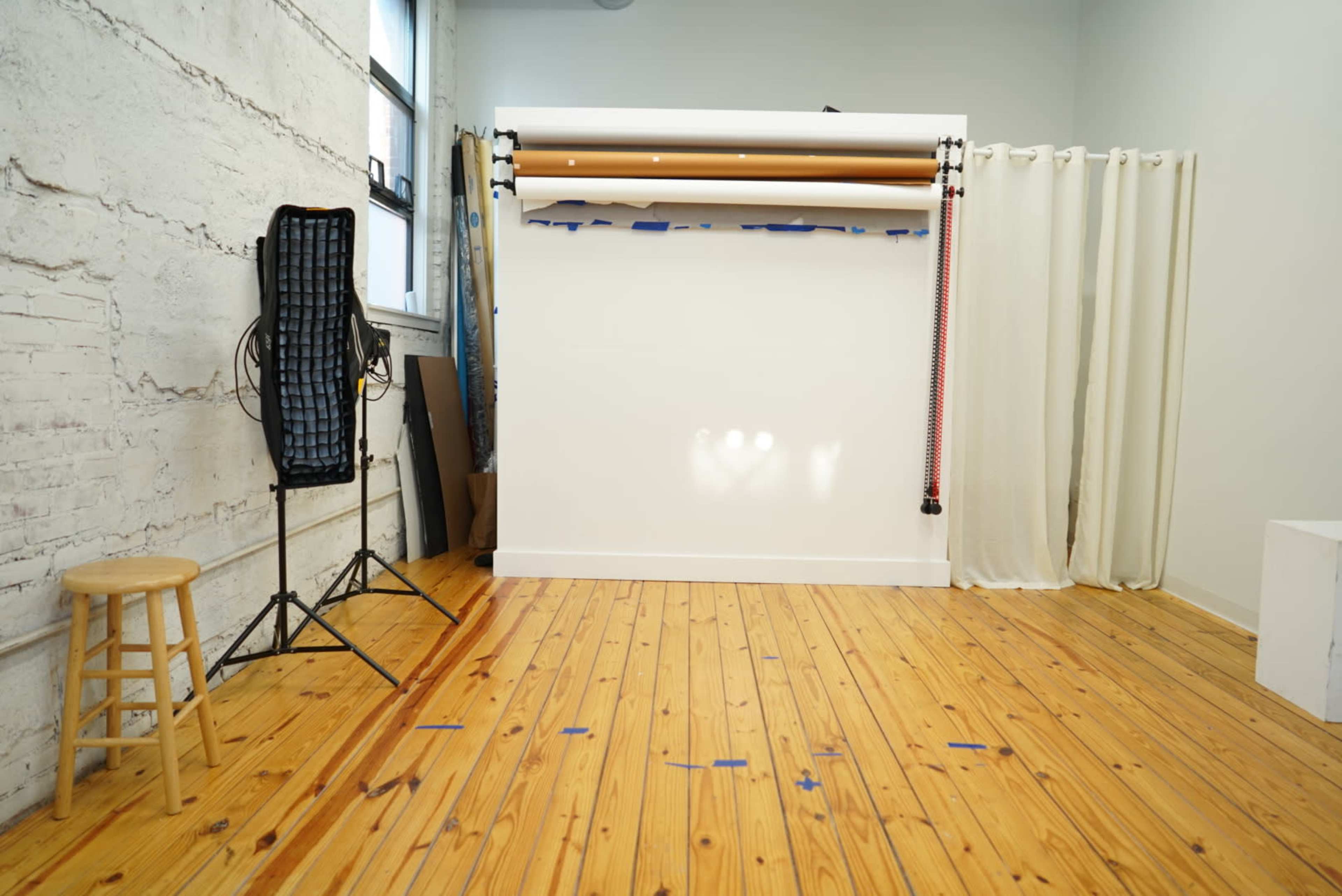 The image shows a photography studio with a wooden floor, a stool, a backdrop stand, and a side curtain.