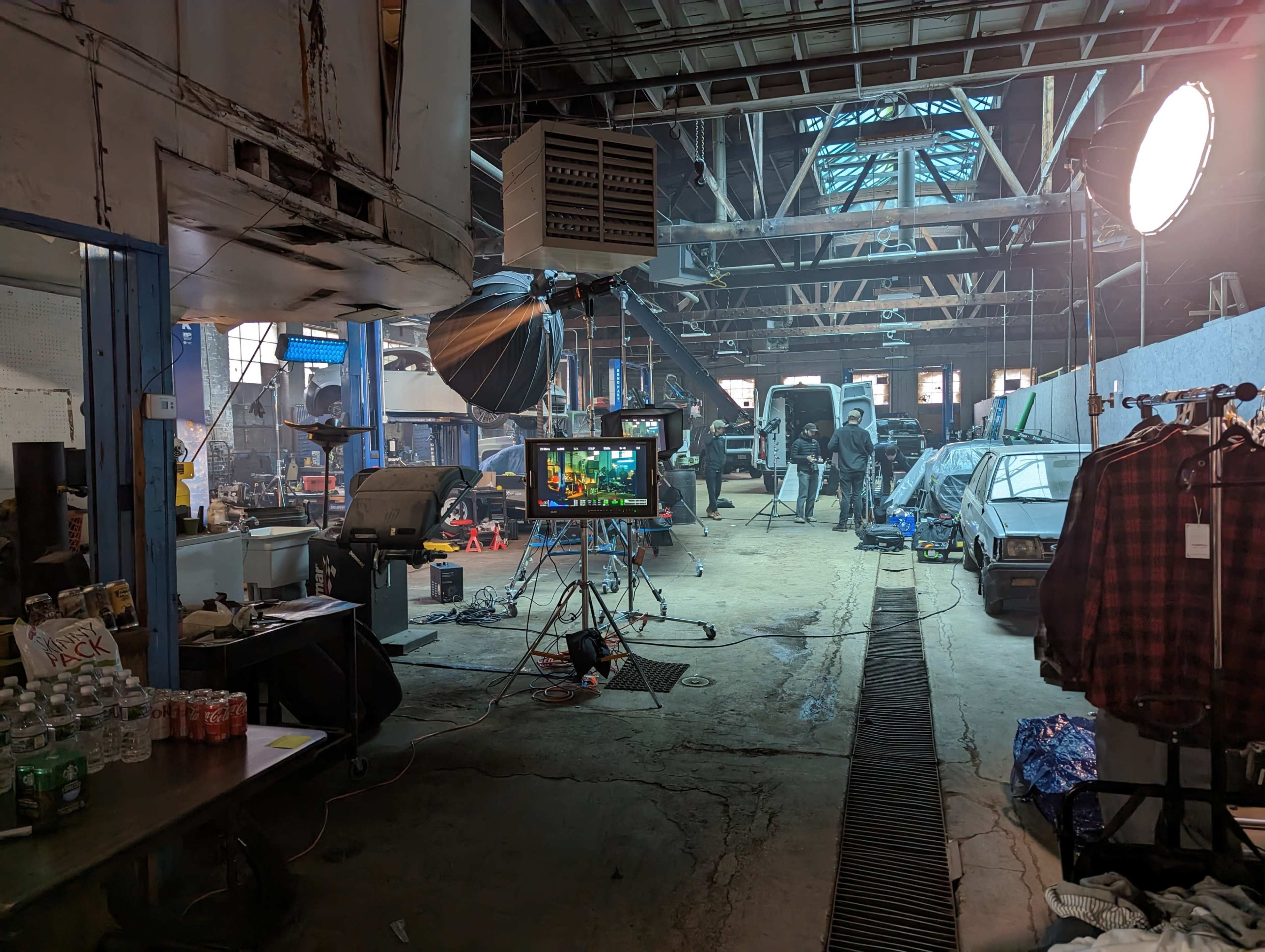A film production set inside a large, industrial space with crew members working around vehicles and equipment.