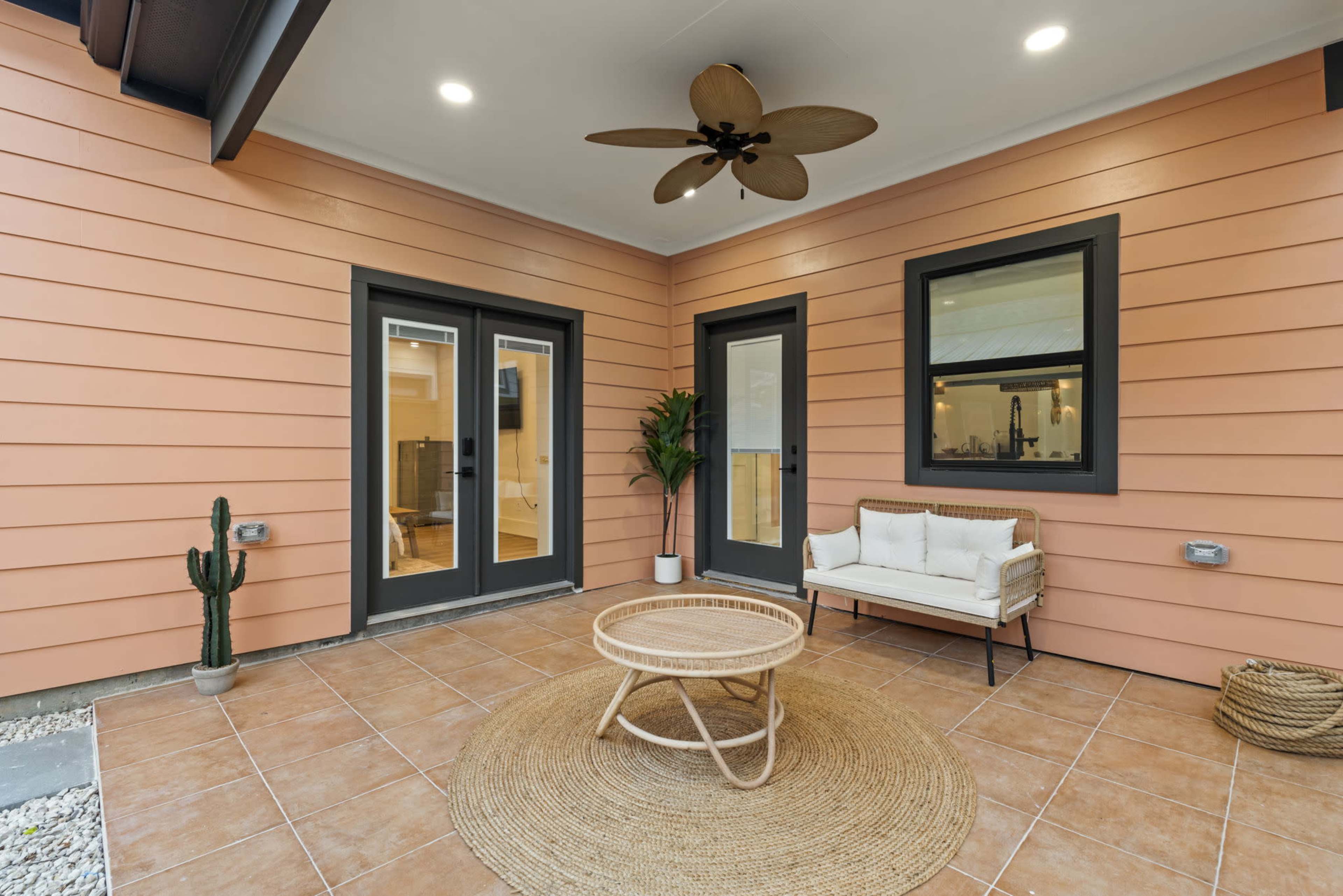 The image features a modern patio with terracotta tiles, a round table in the center, a small sofa against one wall, and two doors leading into the interior of the home.