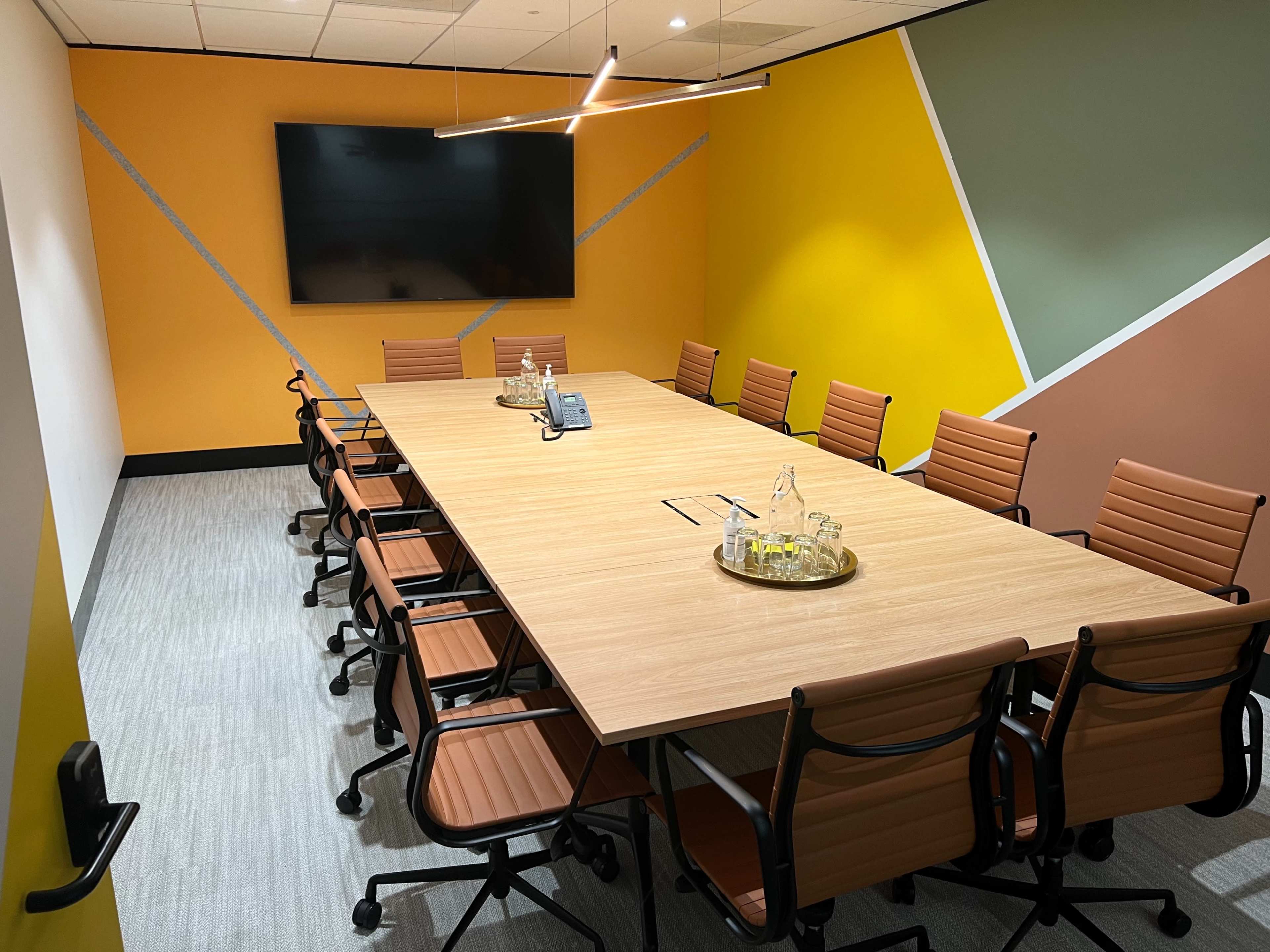 A large conference room is equipped with a rectangular wooden table and ergonomic chairs, featuring a wall-mounted screen and colorful geometric designs.