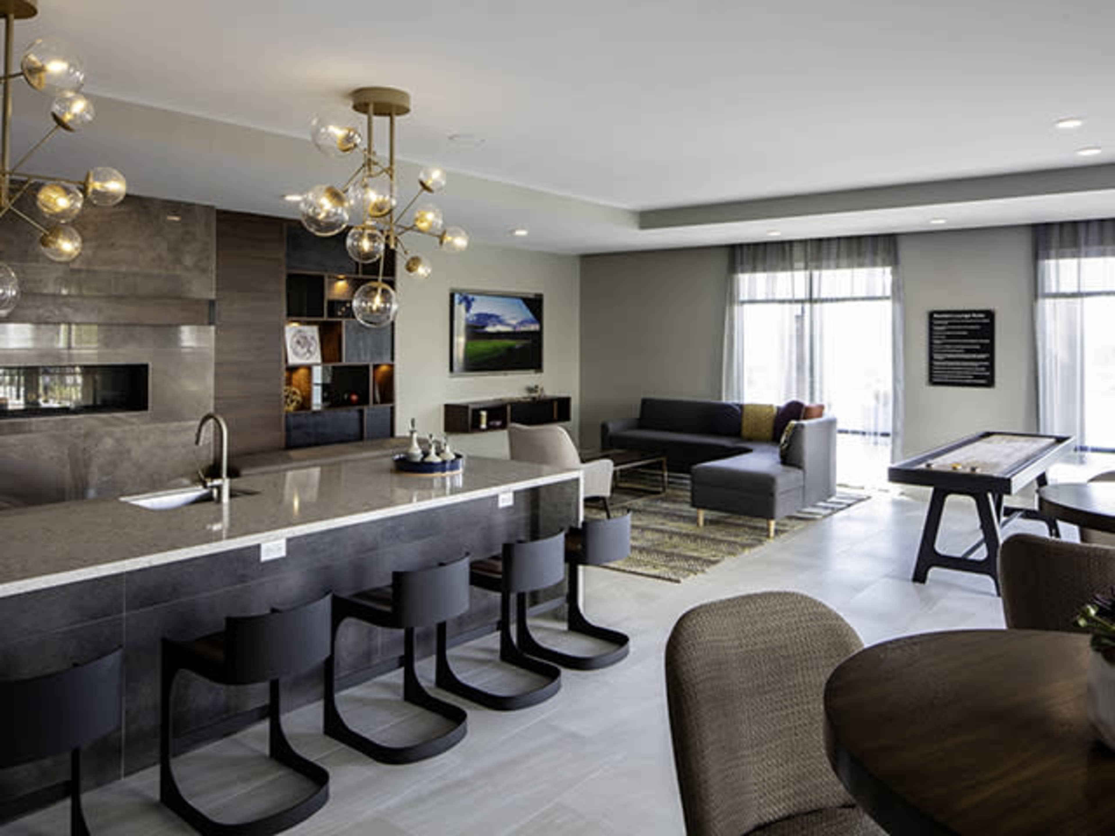 The image shows a modern lounge area featuring a kitchen island with bar stools, a seating area with a gray sofa, and large windows allowing natural light.