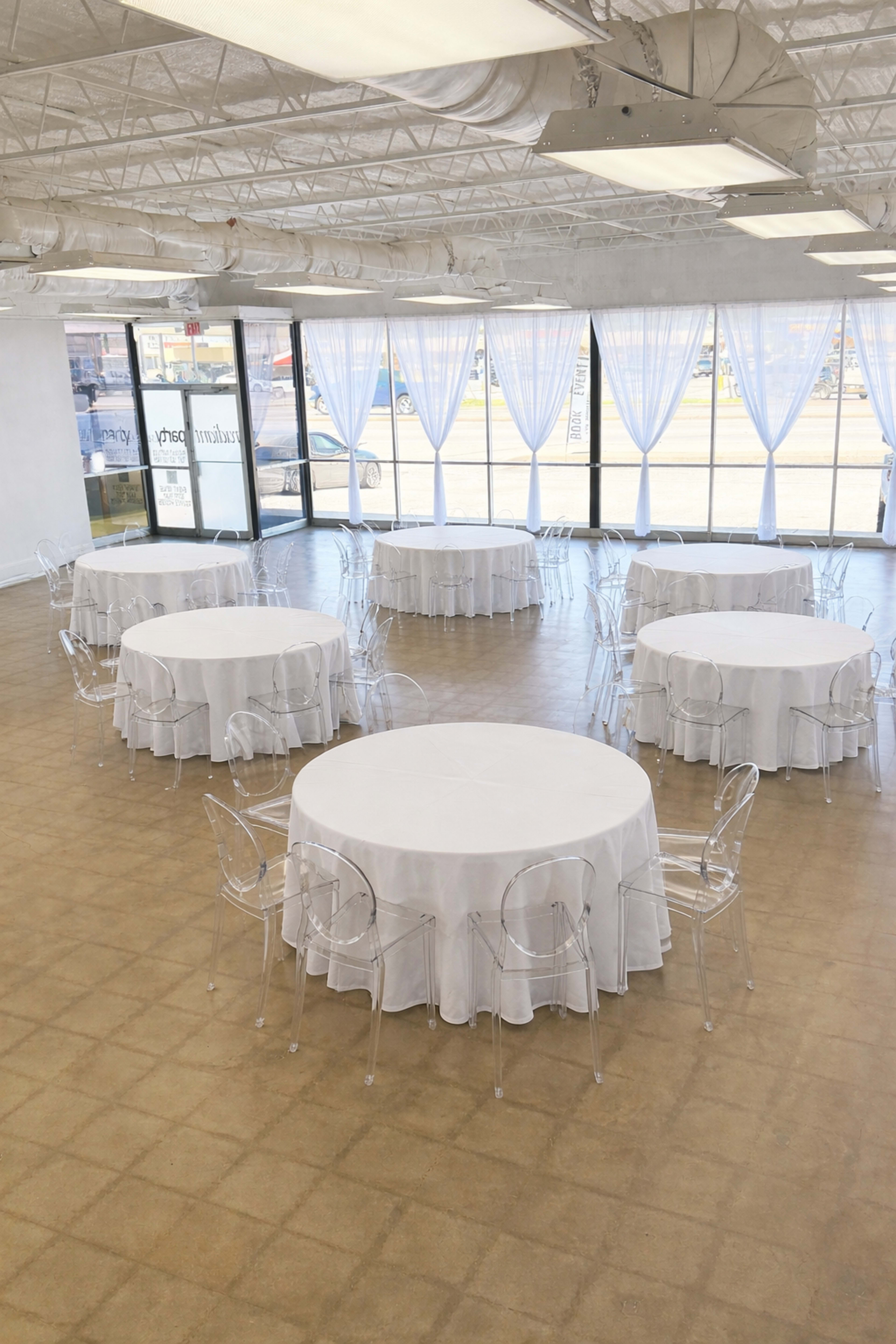 The image shows a spacious event room with several round tables covered in white tablecloths and surrounded by clear acrylic chairs.