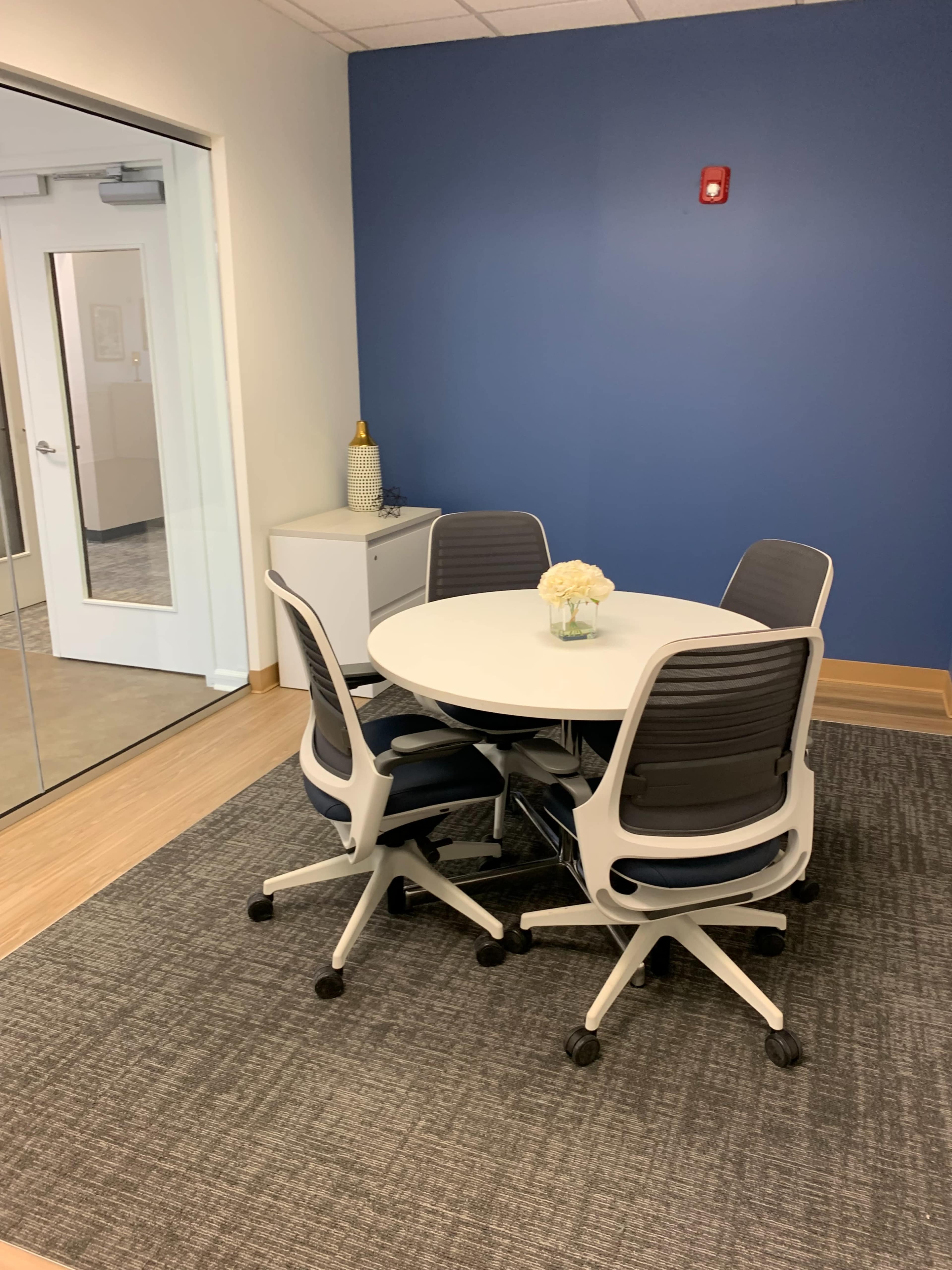 A round table surrounded by four chairs is set in a small meeting room with a blue accent wall and a decorative vase in the center.