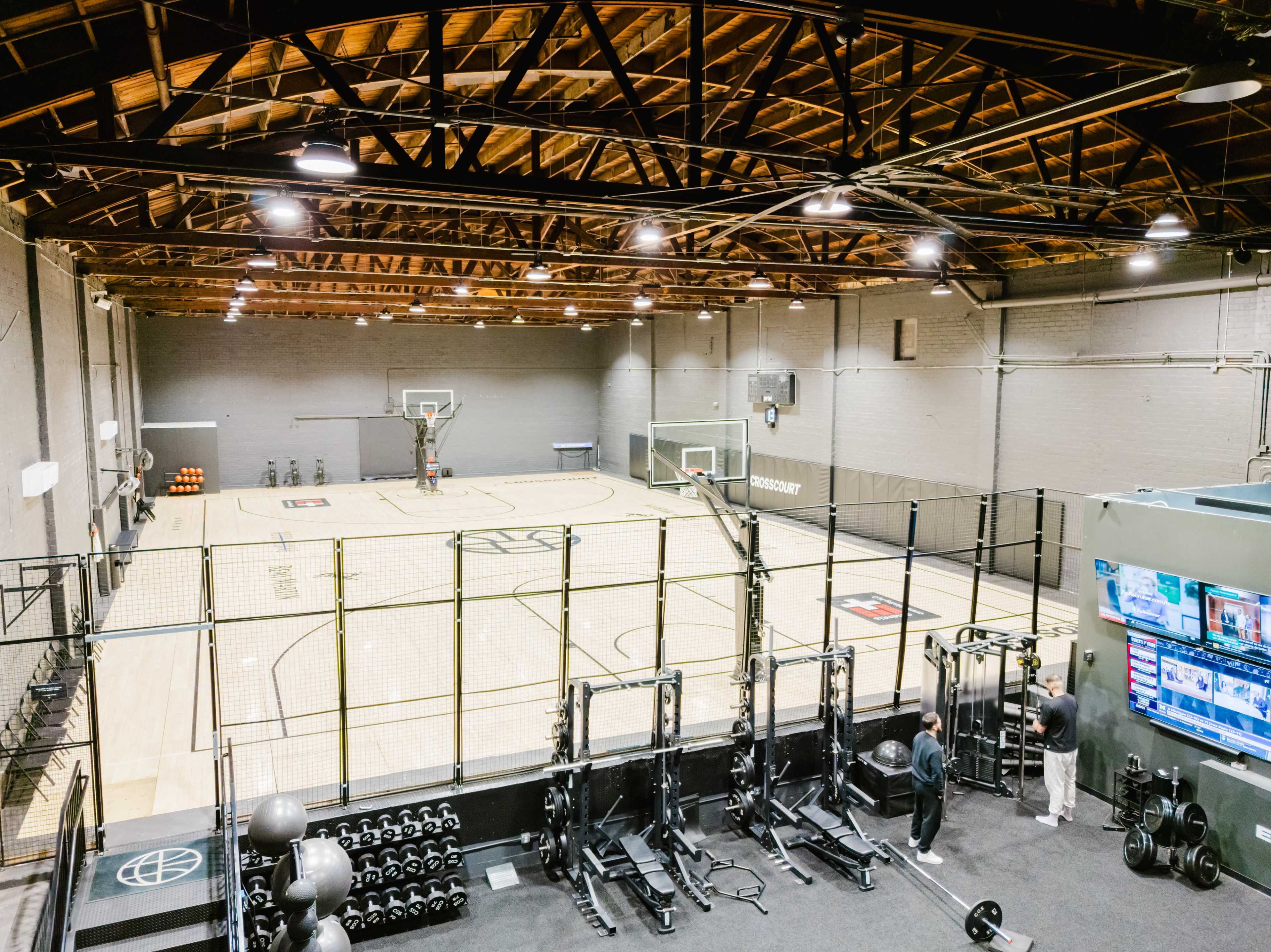 The image shows an indoor basketball court with two hoops, surrounded by a modern workout area featuring weightlifting equipment and screens displaying information.