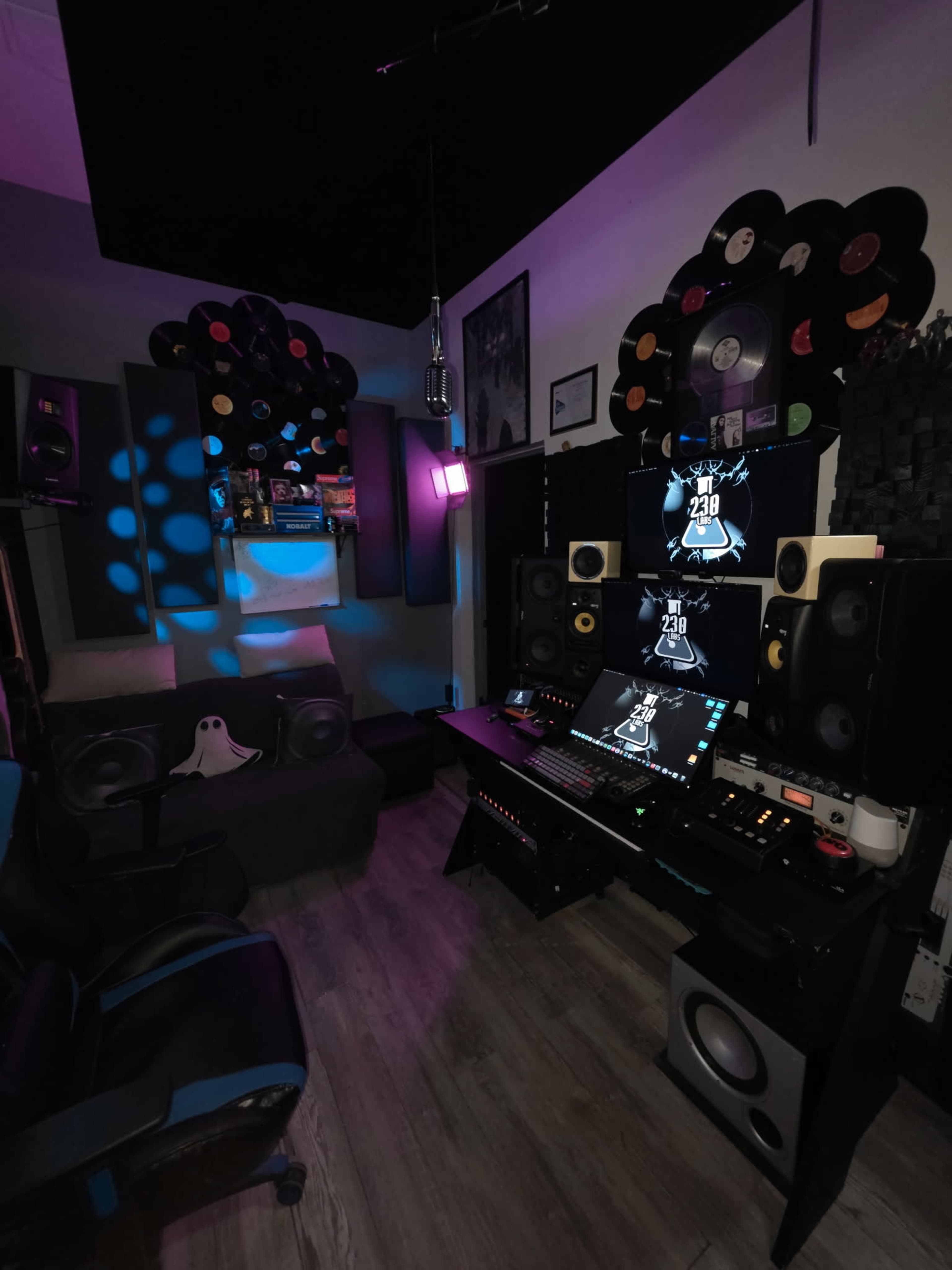 The image shows a music studio featuring a desk with computer monitors, various speakers, and a cozy seating area adorned with vinyl records on the walls.