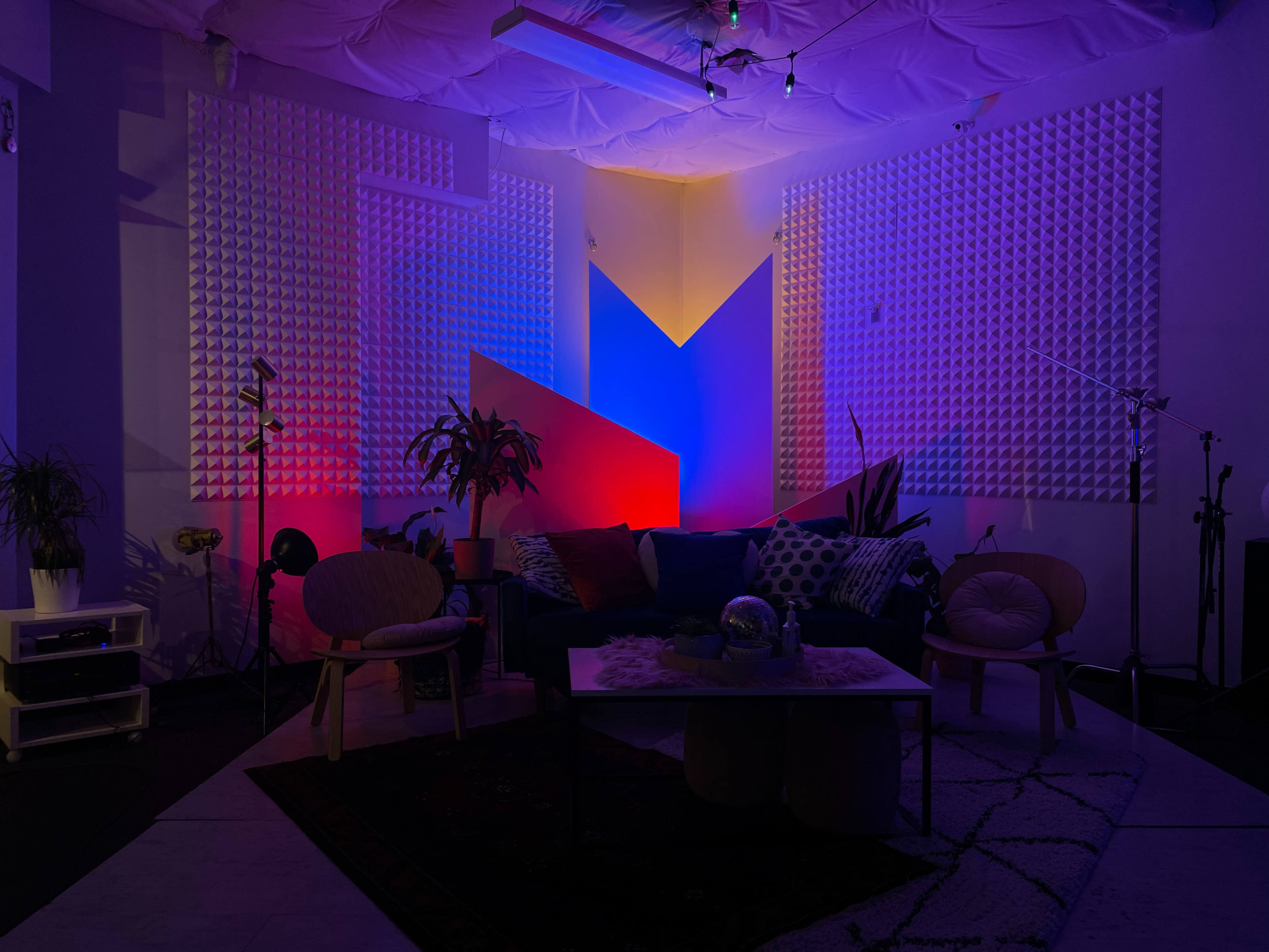 A dimly lit lounge area with geometric wall decor, colorful lighting, a couch adorned with pillows, and scattered furniture.
