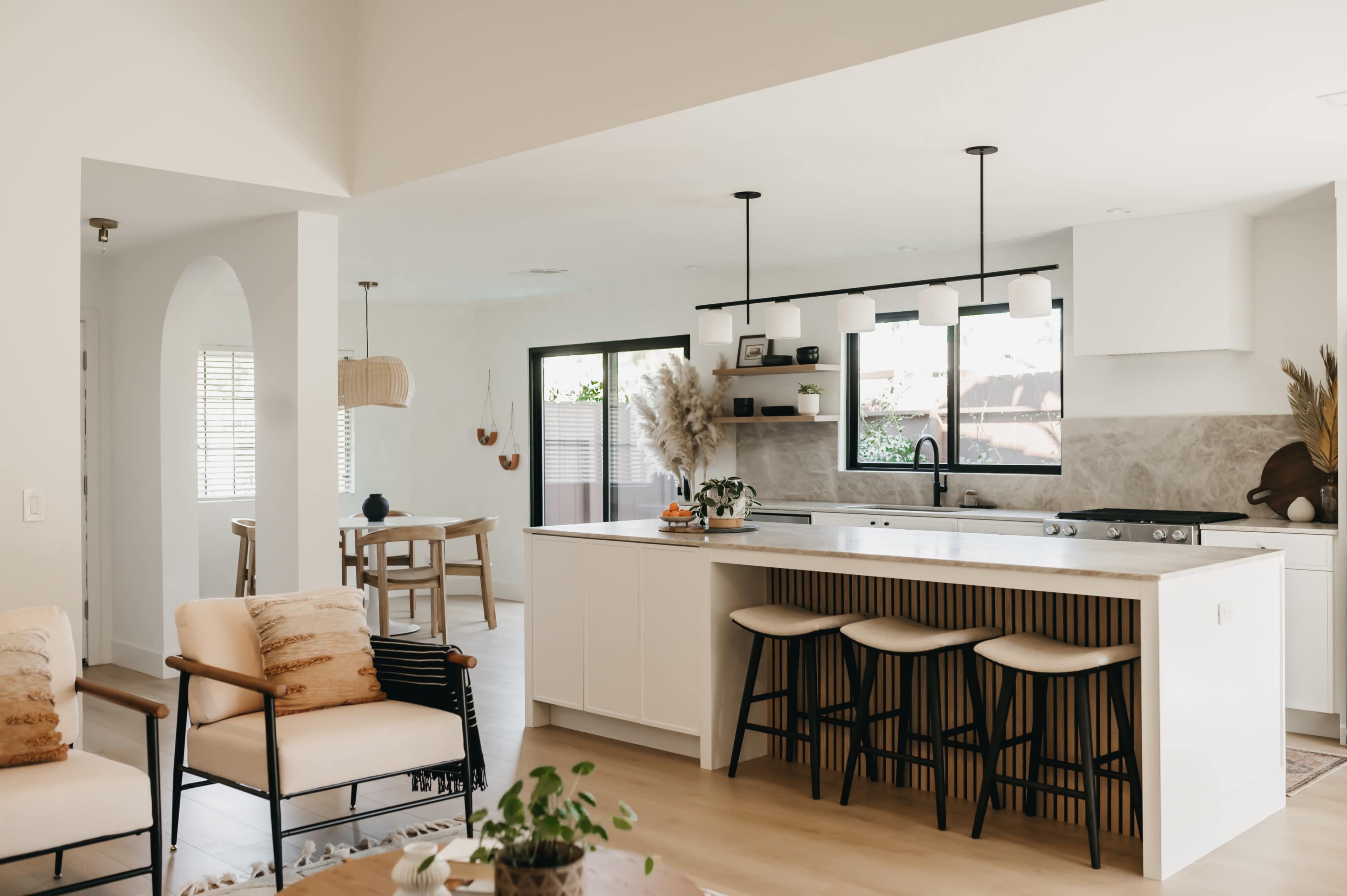 A modern kitchen with white cabinetry, a breakfast bar with three stools, and an adjacent dining area featuring a wooden table and minimal decor.