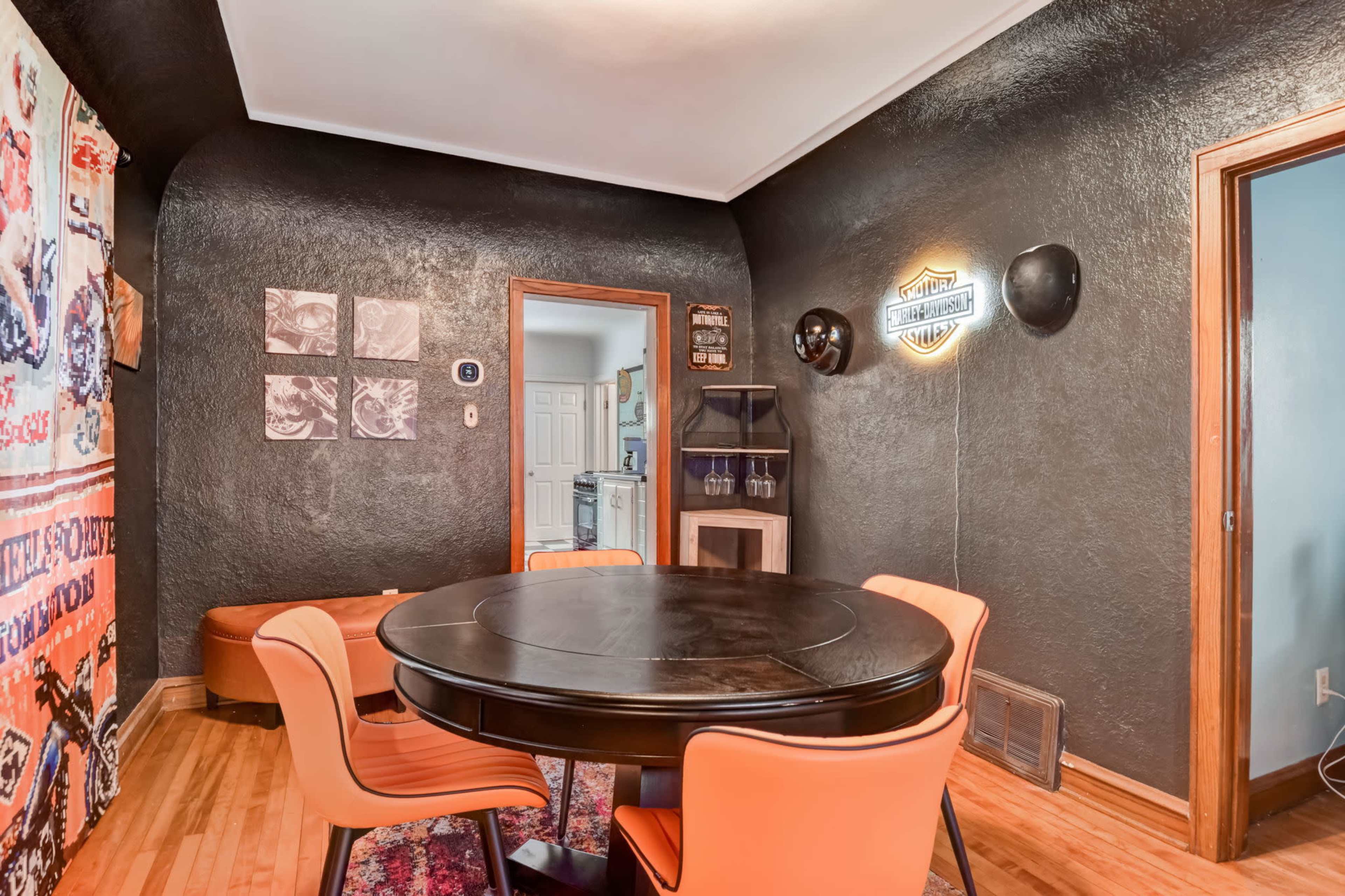 A circular black dining table is surrounded by orange chairs in a room with dark walls and a decorative wall hanging.