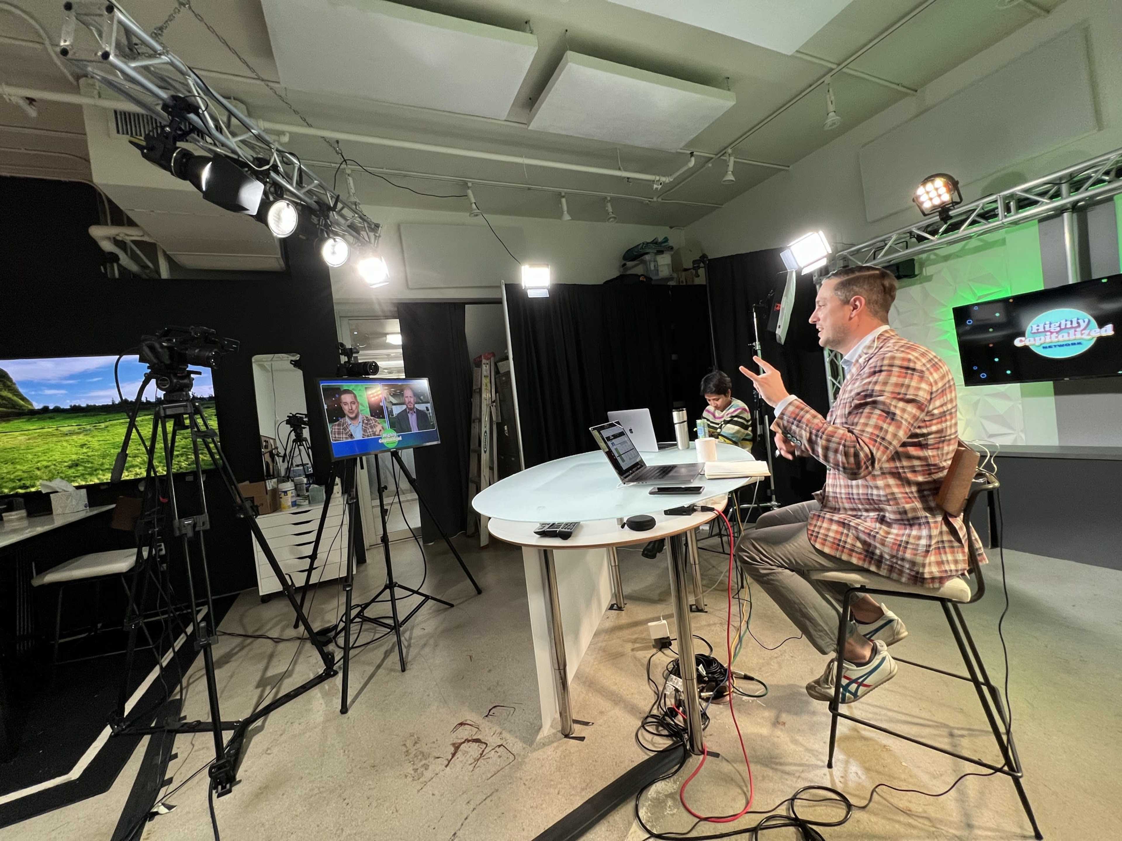 A man in a plaid jacket gestures while seated at a table in a studio, with cameras, lights, and a screen displaying two video calls in the background.