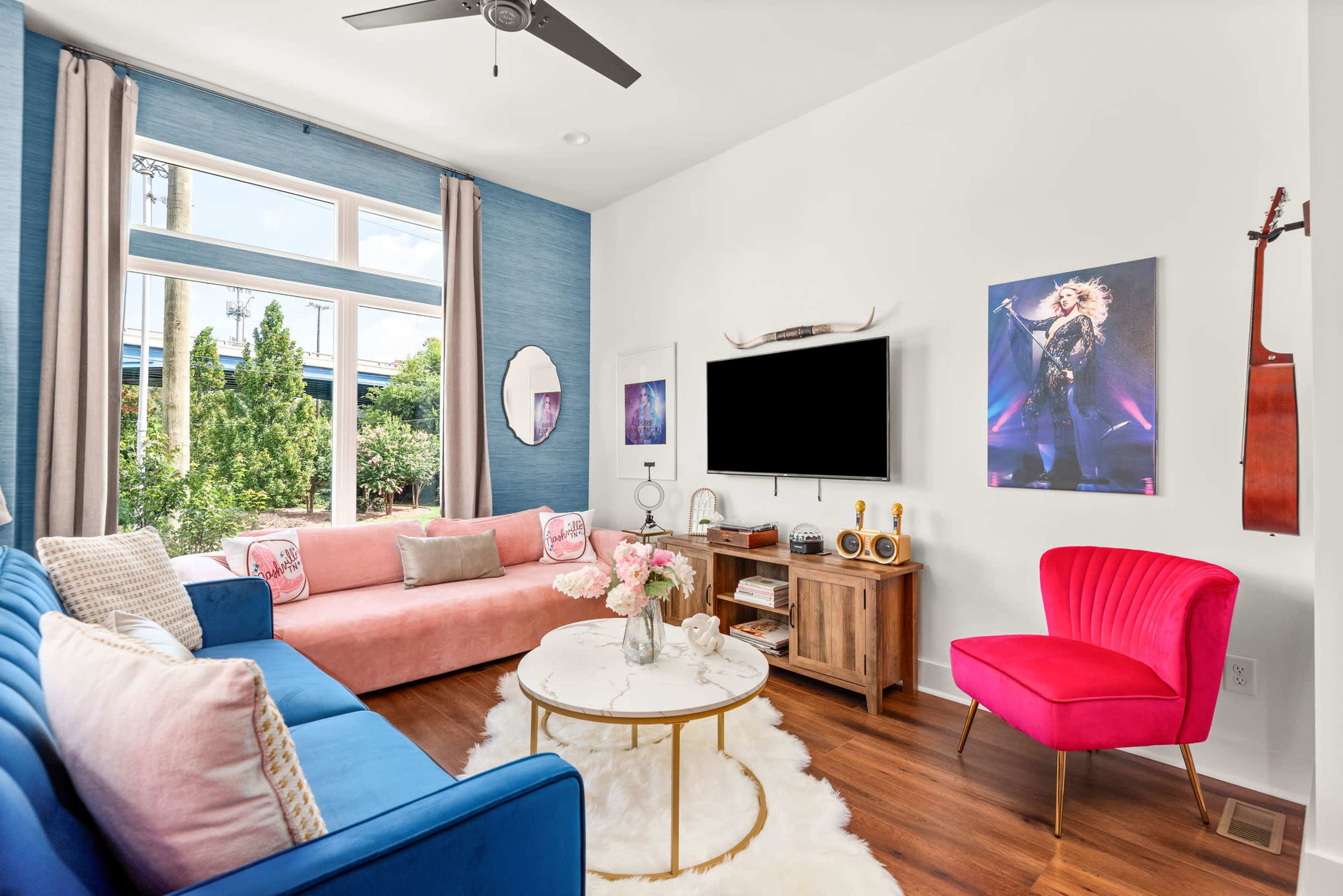 The image shows a contemporary living room featuring a pink sofa, a blue couch, a round coffee table, and a TV mounted on the wall, with a large window providing natural light.