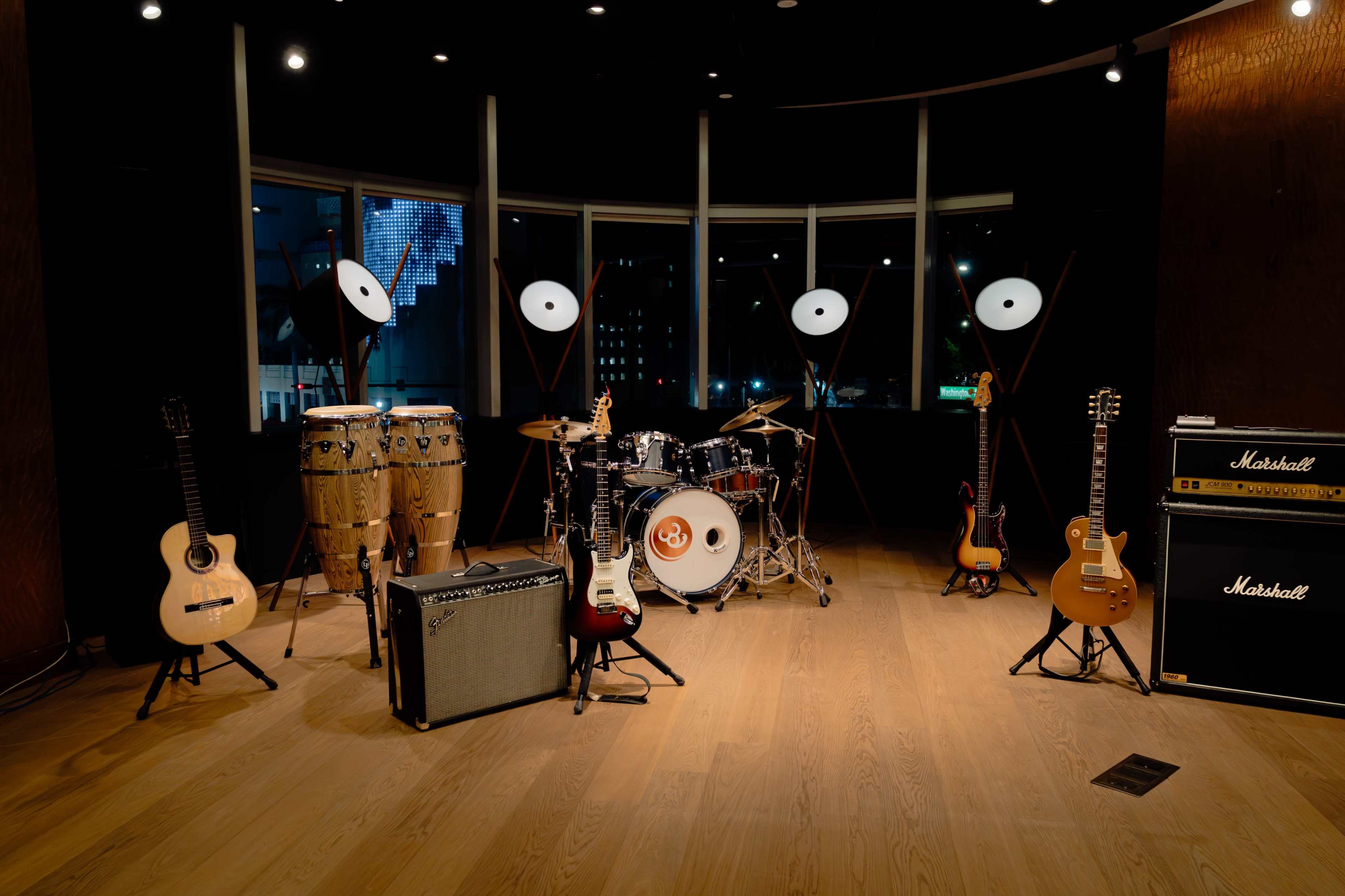 The image shows a music setup featuring various instruments, including guitars, drums, and amplifiers, arranged in a well-lit room with large windows.