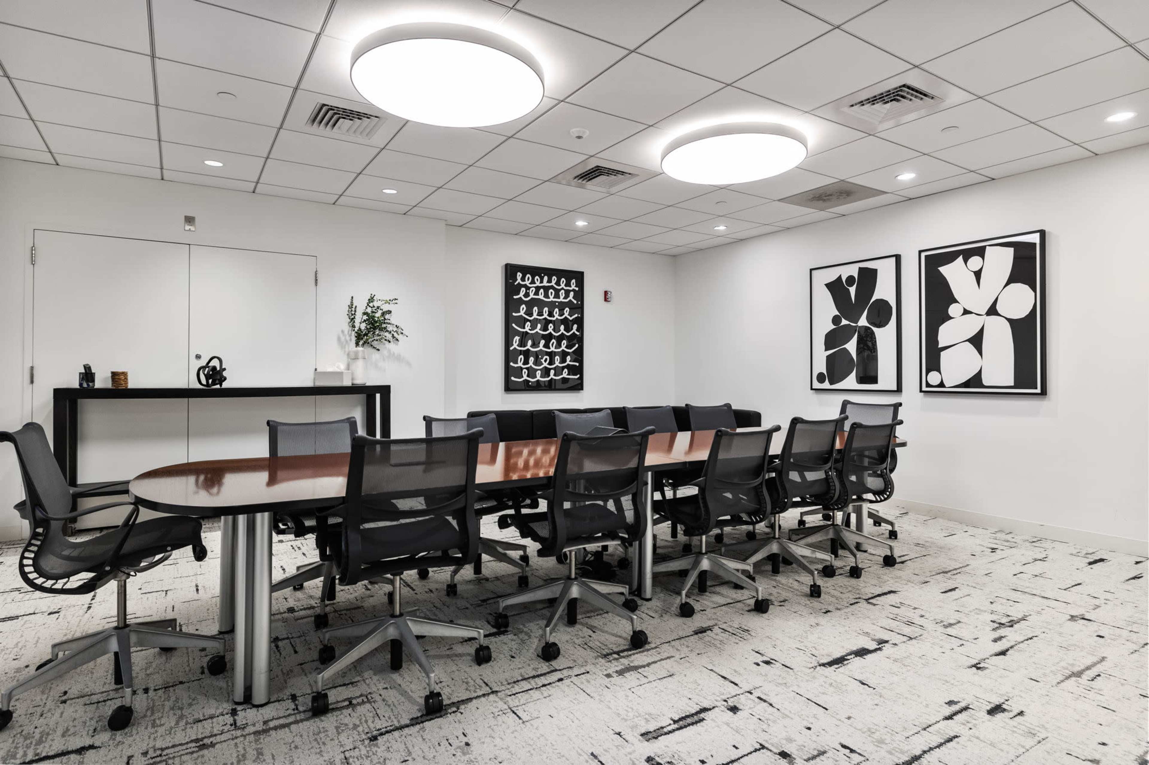 The image shows a modern conference room with a long table surrounded by ergonomic chairs, featuring abstract artwork on the walls.