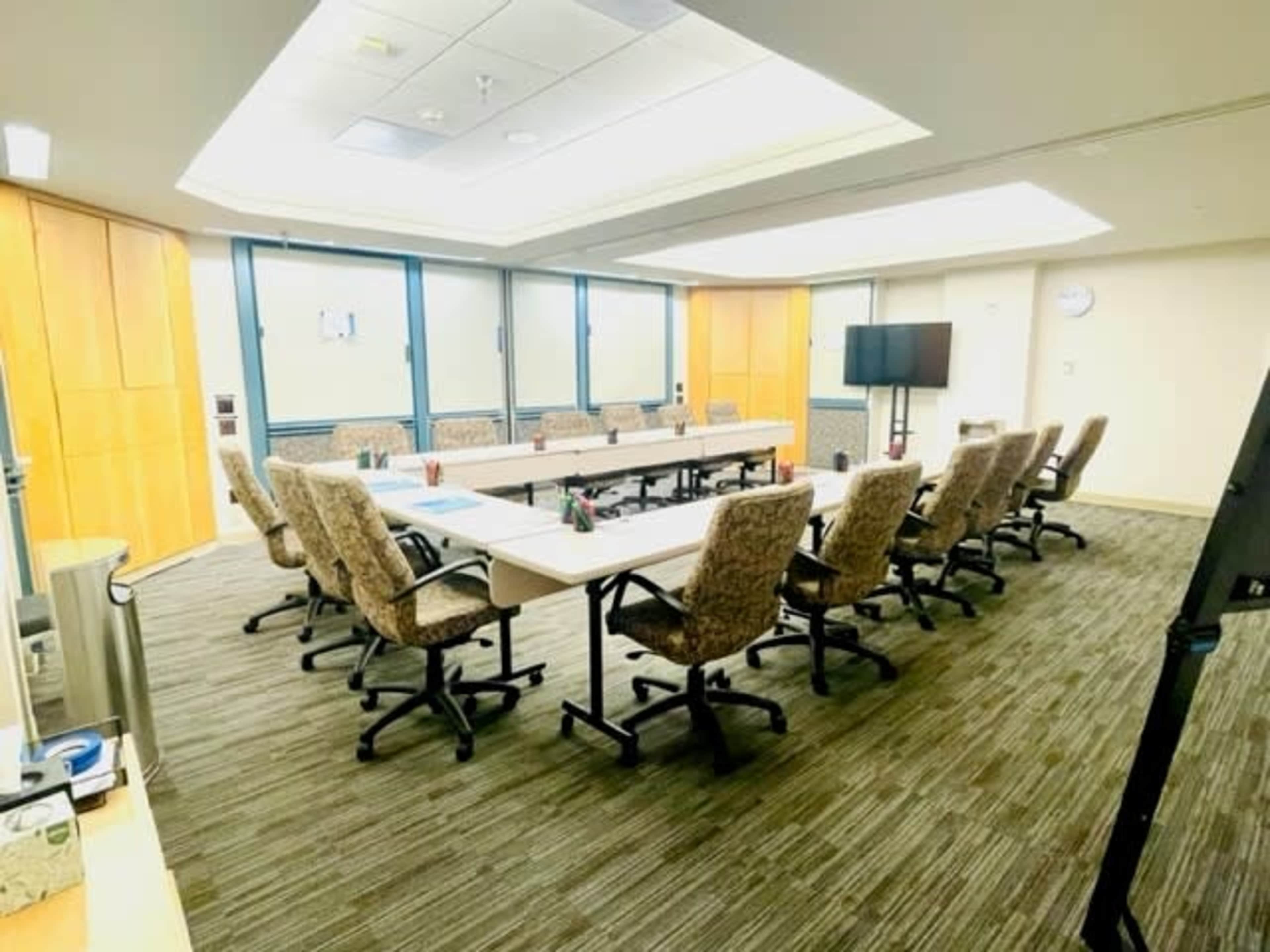 A conference room with a long table surrounded by rolling chairs, featuring a television and large windows.
