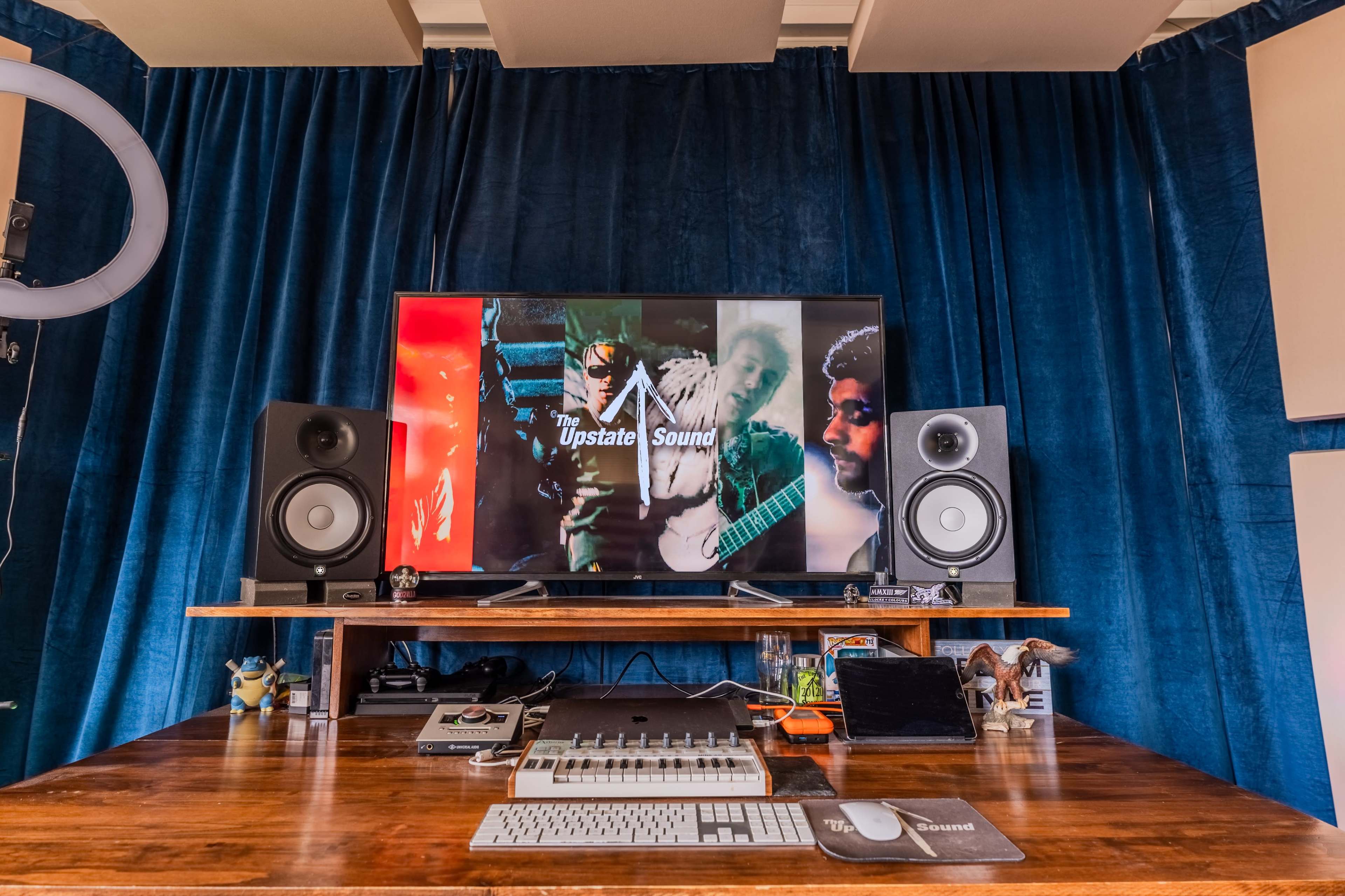 A wooden desk with audio equipment and a large television displaying a logo and images of musicians is set against a blue velvet curtain.