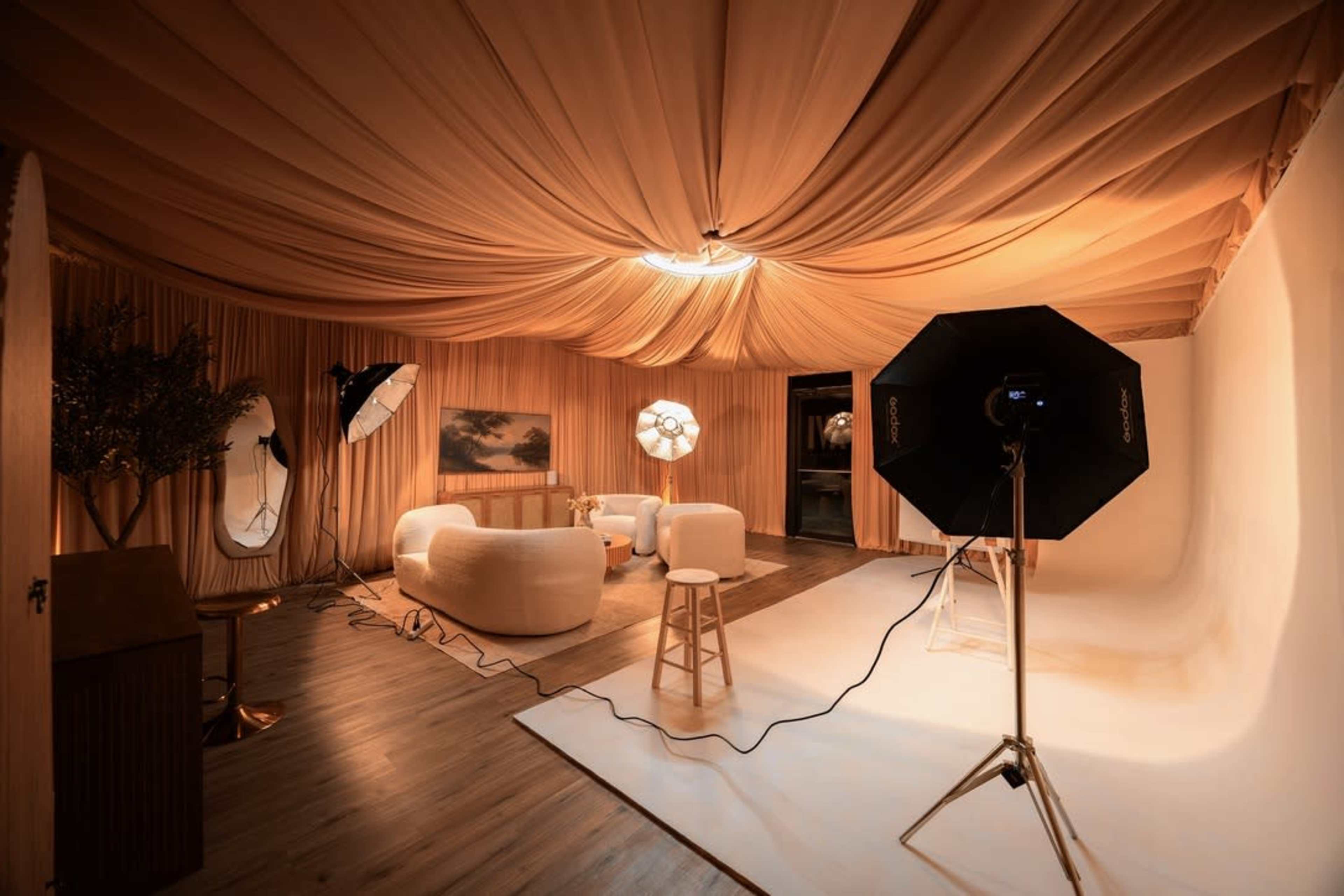 The image shows a photography studio with a softly draped ceiling, a seating area featuring white furniture, and professional lighting equipment set up for a shoot.