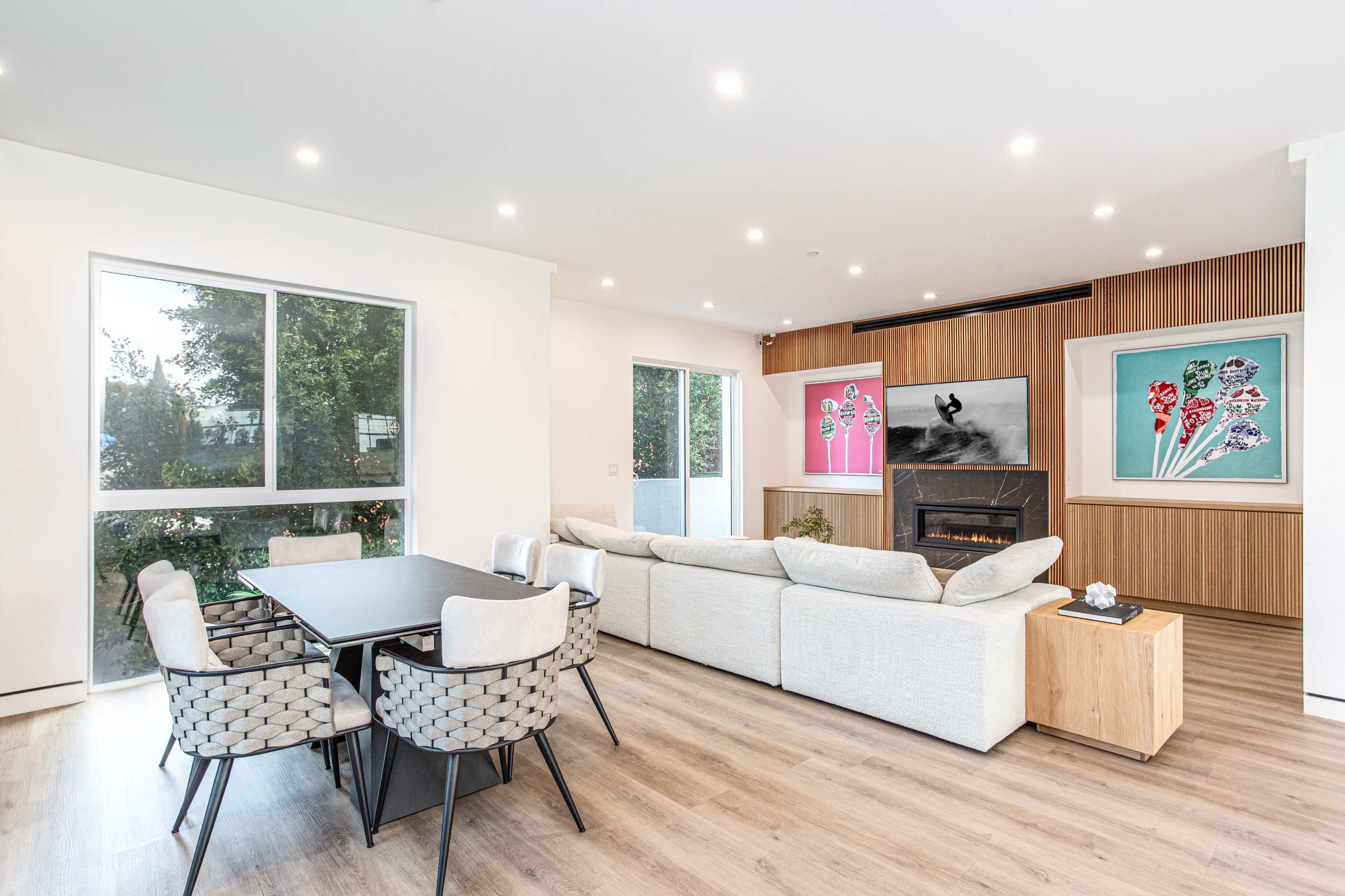A modern living space with a dining area featuring a black table and patterned chairs, a light-colored sofa, a wall-mounted fireplace, and colorful artwork above a wooden panel.