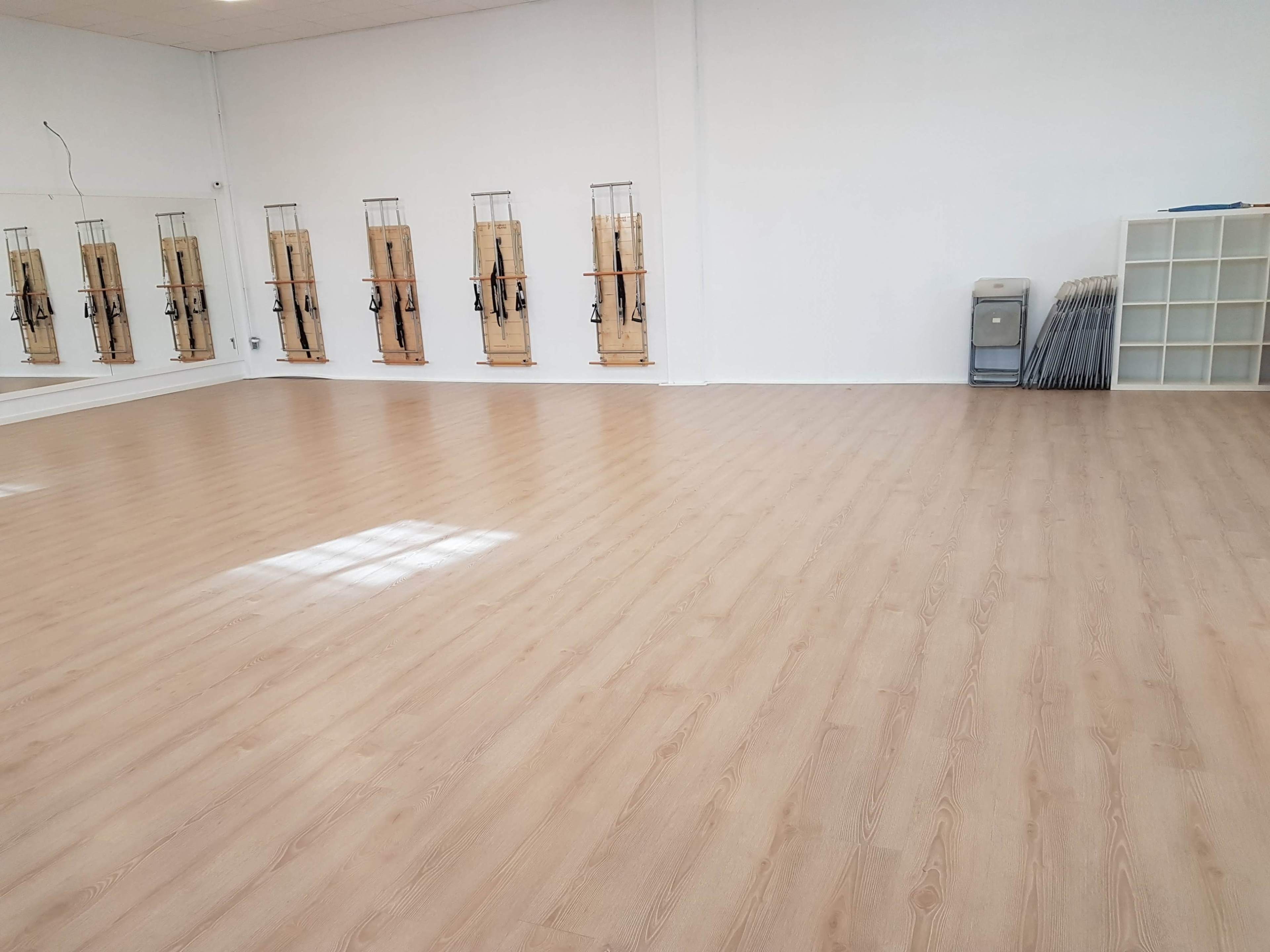 An empty fitness studio with wooden flooring, mirrors on one wall, and equipment racks along another wall.