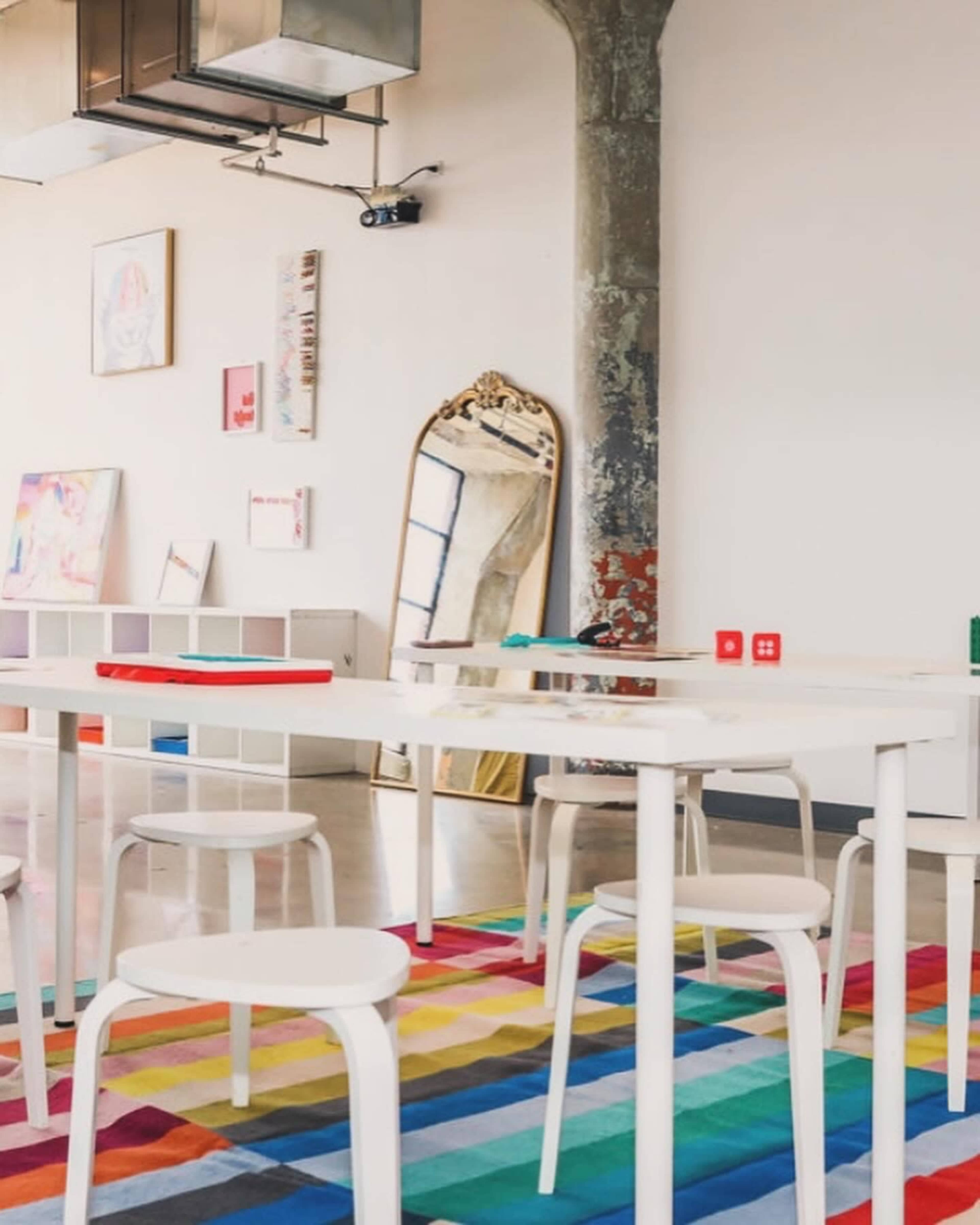 The scene features a brightly colored, patterned rug on a polished floor, with white tables and stools arranged around a large ornate mirror against a light-colored wall.