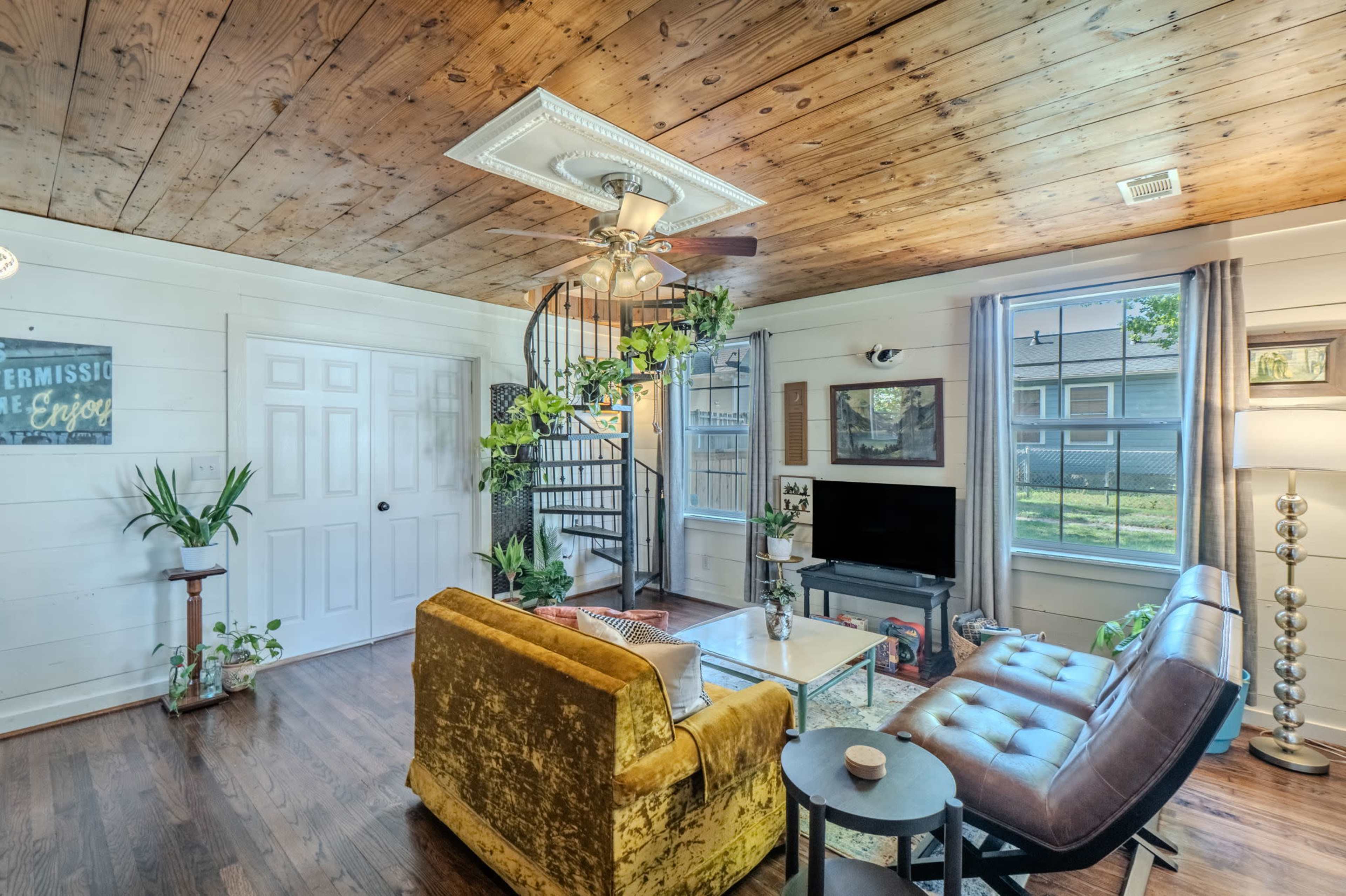 The living room features a spiral staircase, a television, and a seating area with a yellow couch and a brown chair, surrounded by plants and windows.