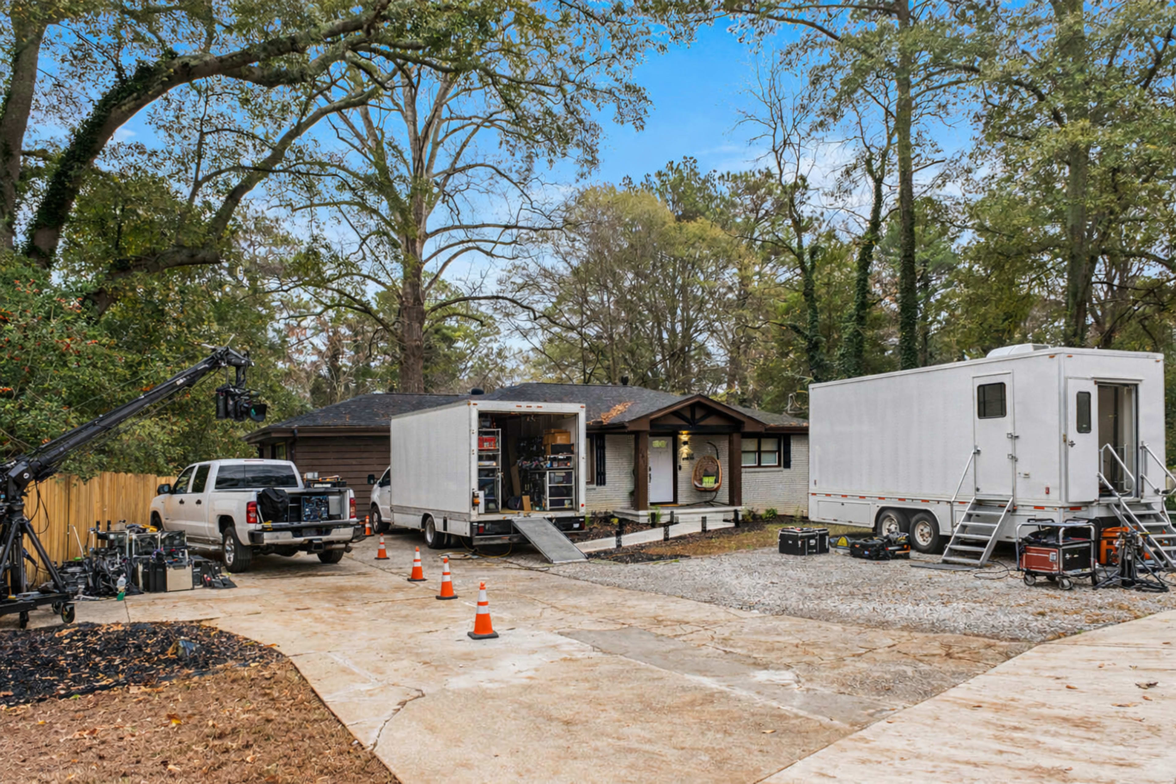 A film crew is set up outside a house, with equipment trucks and a crane positioned on a gravel driveway.