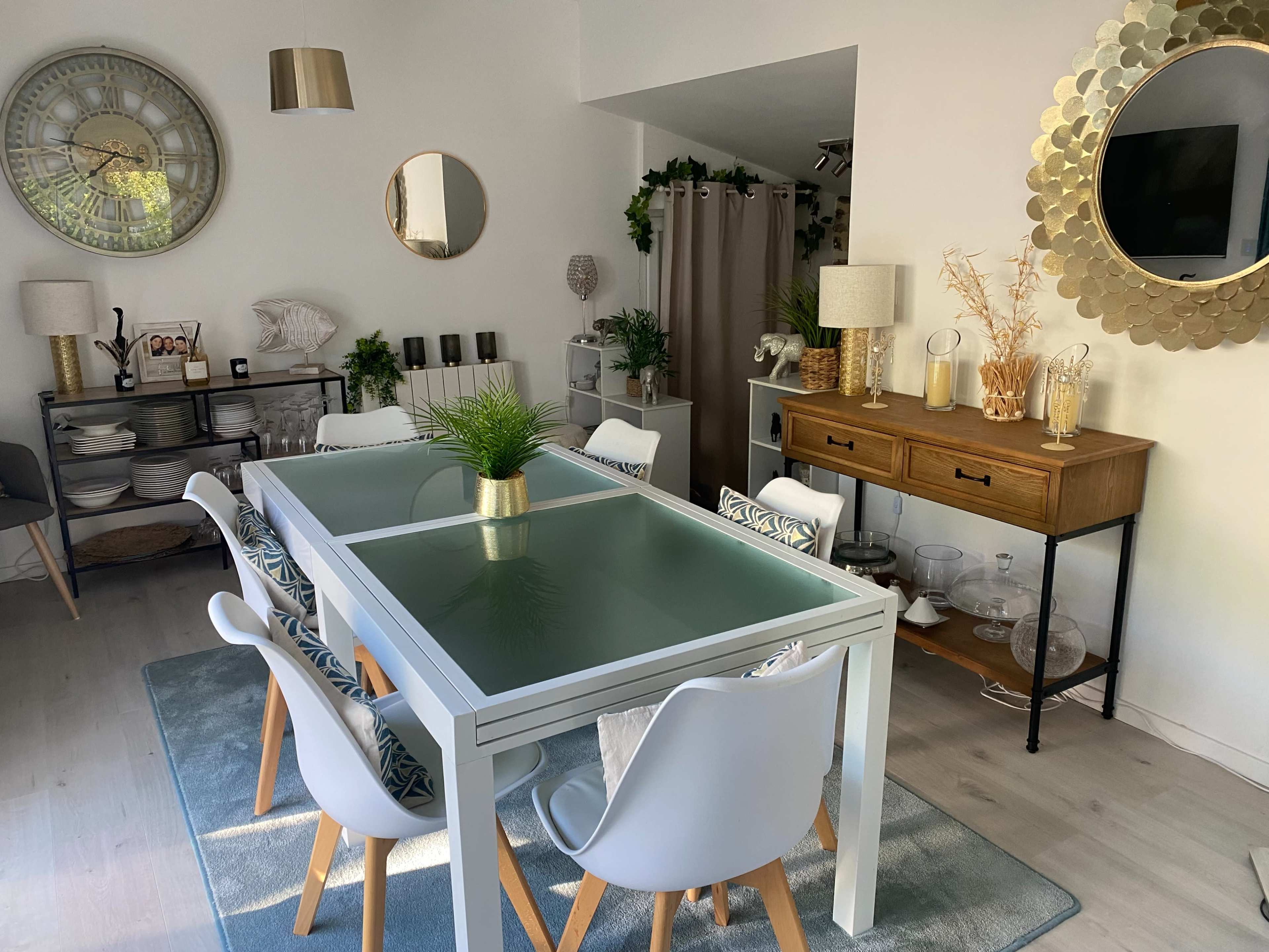 A modern dining area features a glass-topped table surrounded by white chairs, with decorative elements and a sideboard in a well-lit room.