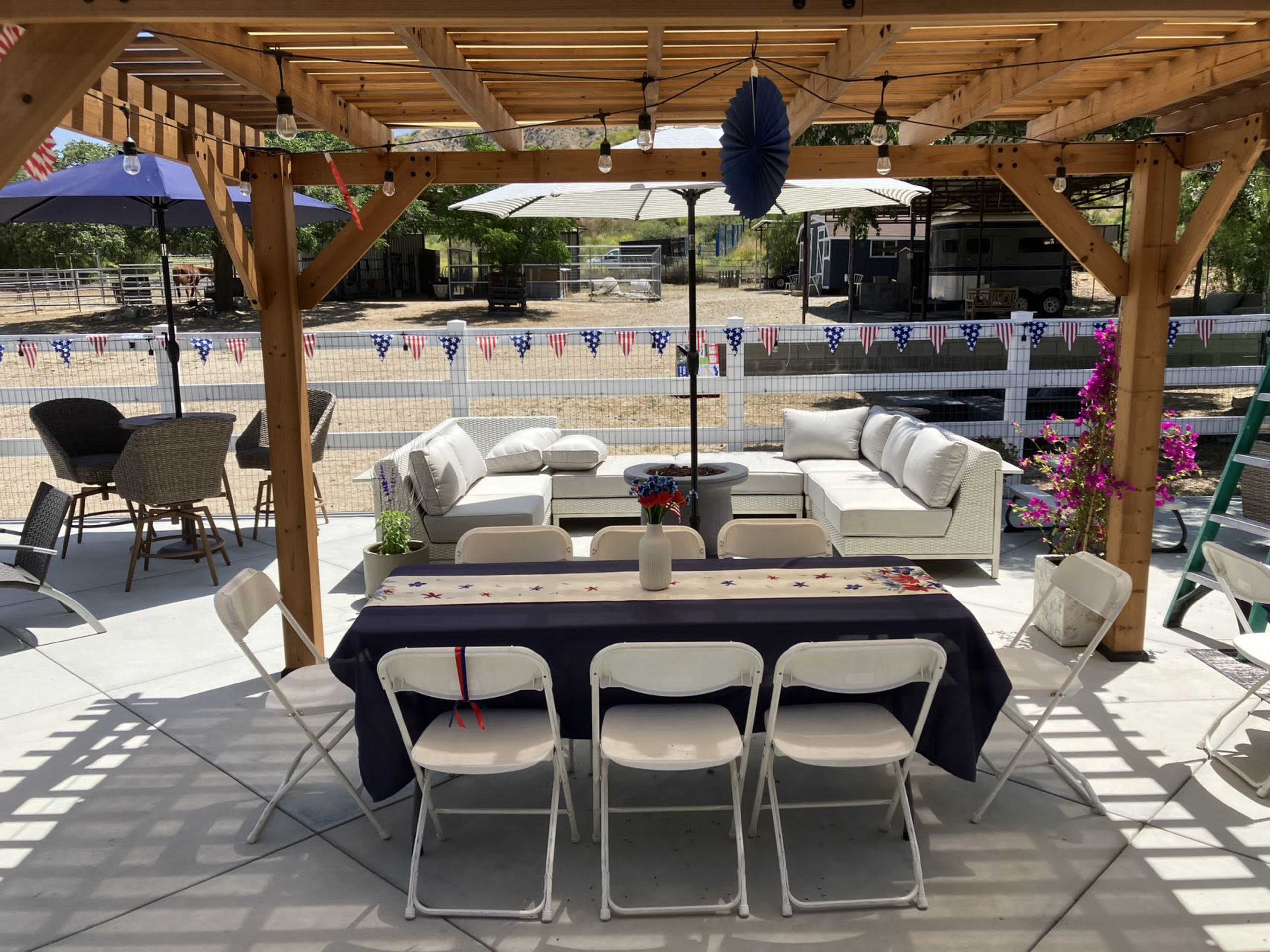 A shaded outdoor patio features a long table set with white chairs, surrounded by a seating area with couches and decorative elements.