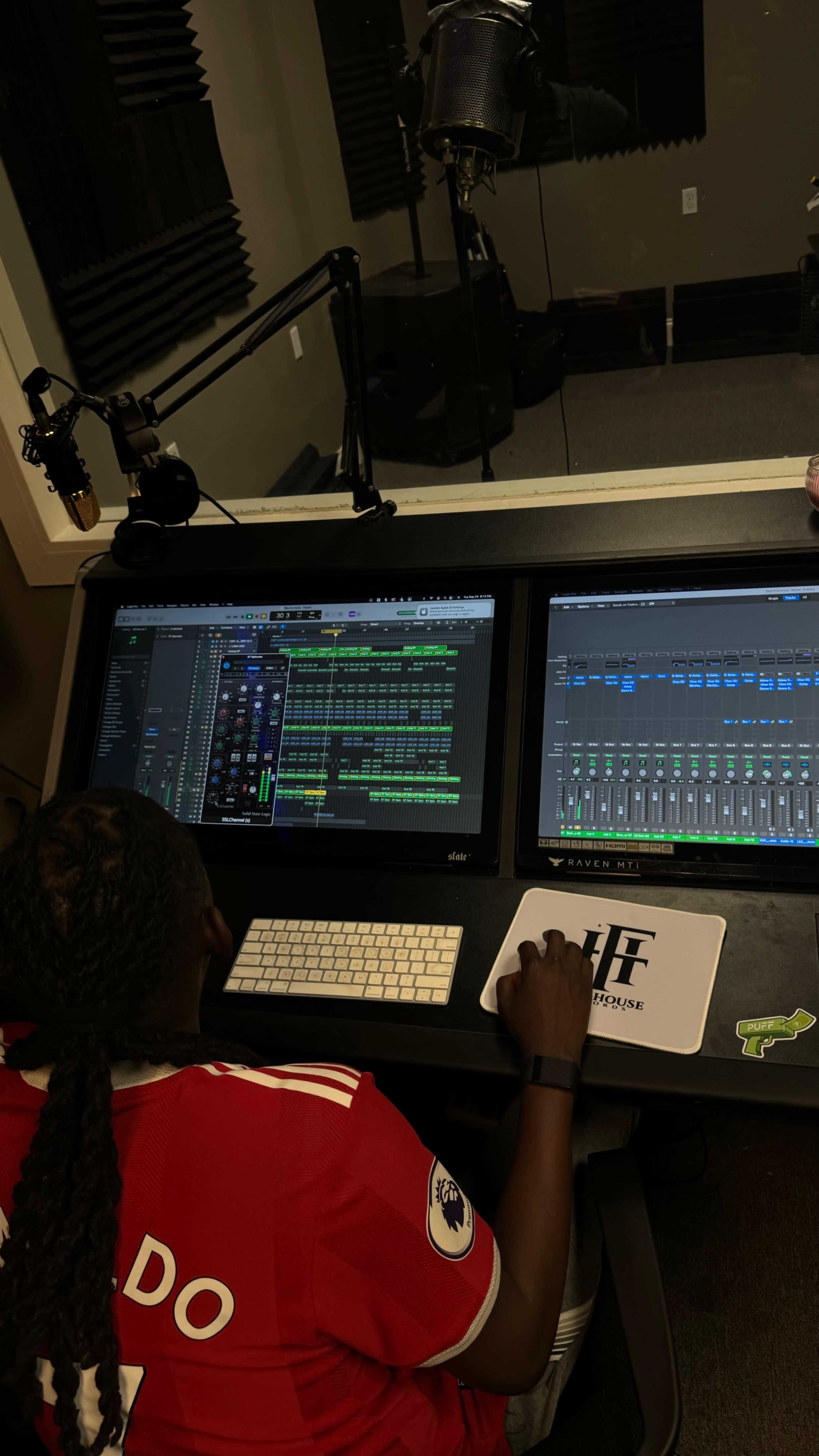 A person is working at a dual-screen audio production setup, with music software visible on the monitors and recording equipment in the background.