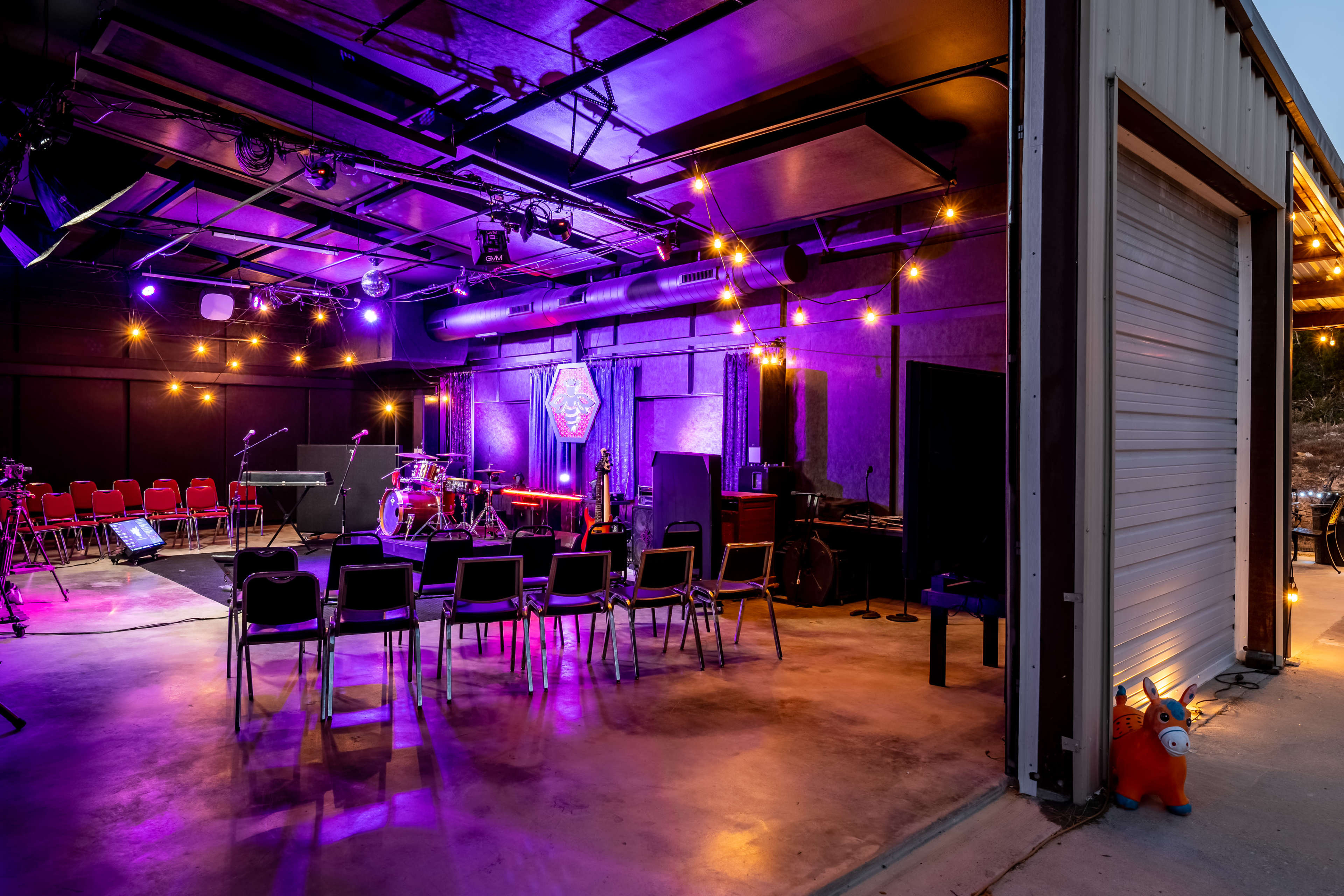 The image shows a modern performance space with stage lighting, musical instruments set up, and rows of chairs arranged for an audience.