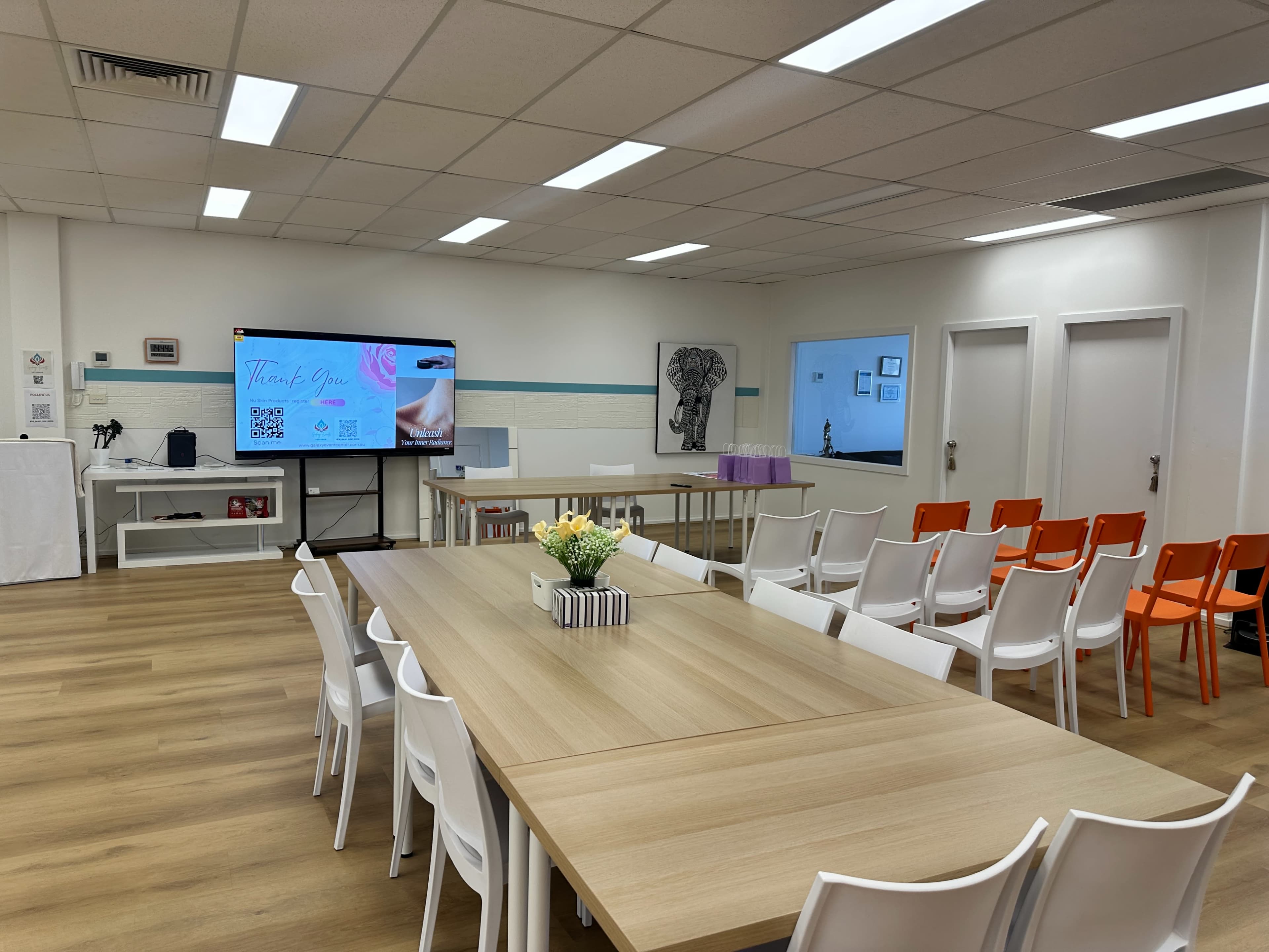 The image shows a spacious meeting room with several tables arranged, chairs set up in rows, and a large screen displaying a presentation.