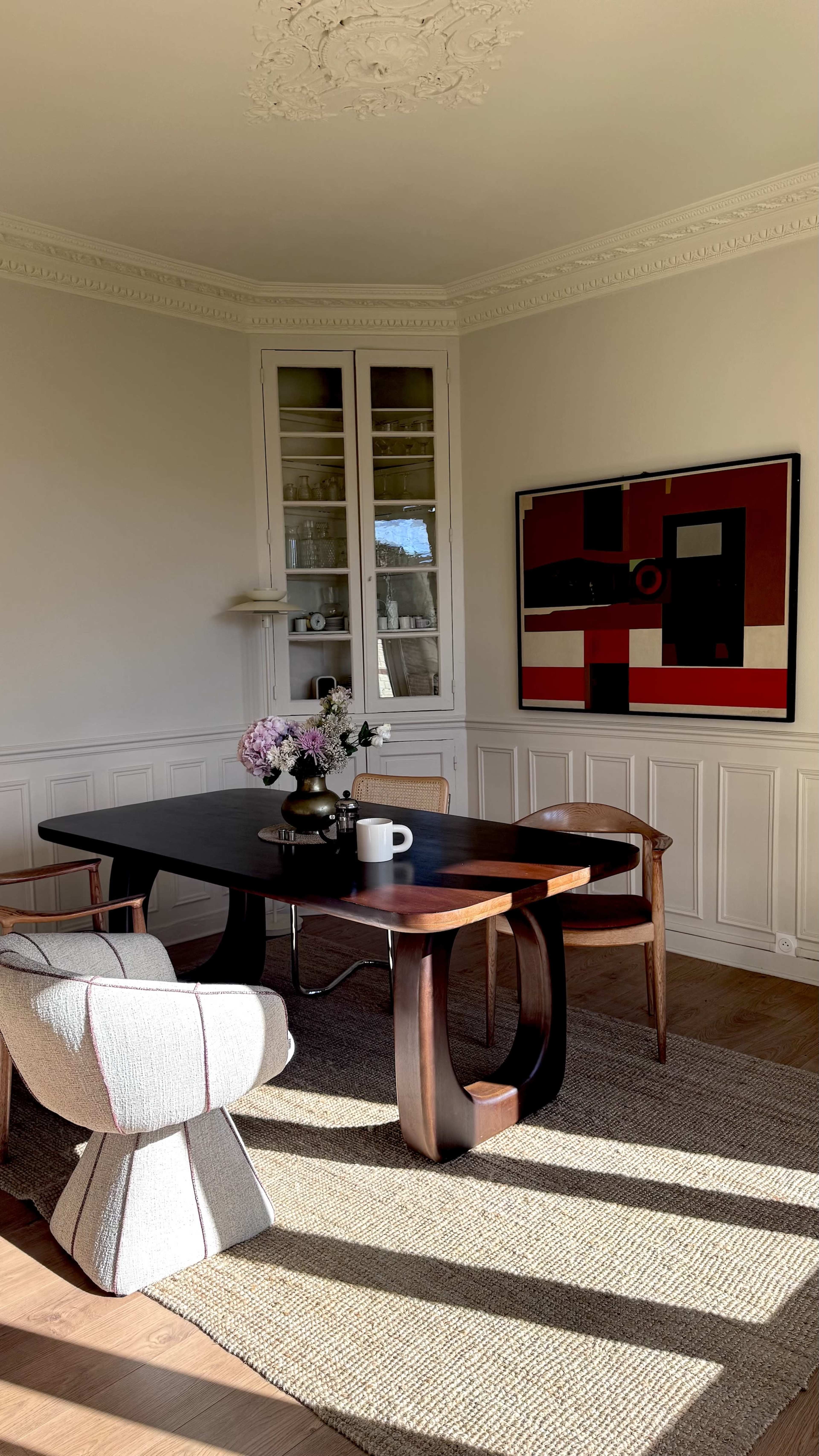 A modern dining area features a dark wooden table surrounded by stylish chairs, a vase of flowers, and a large abstract painting on the wall.