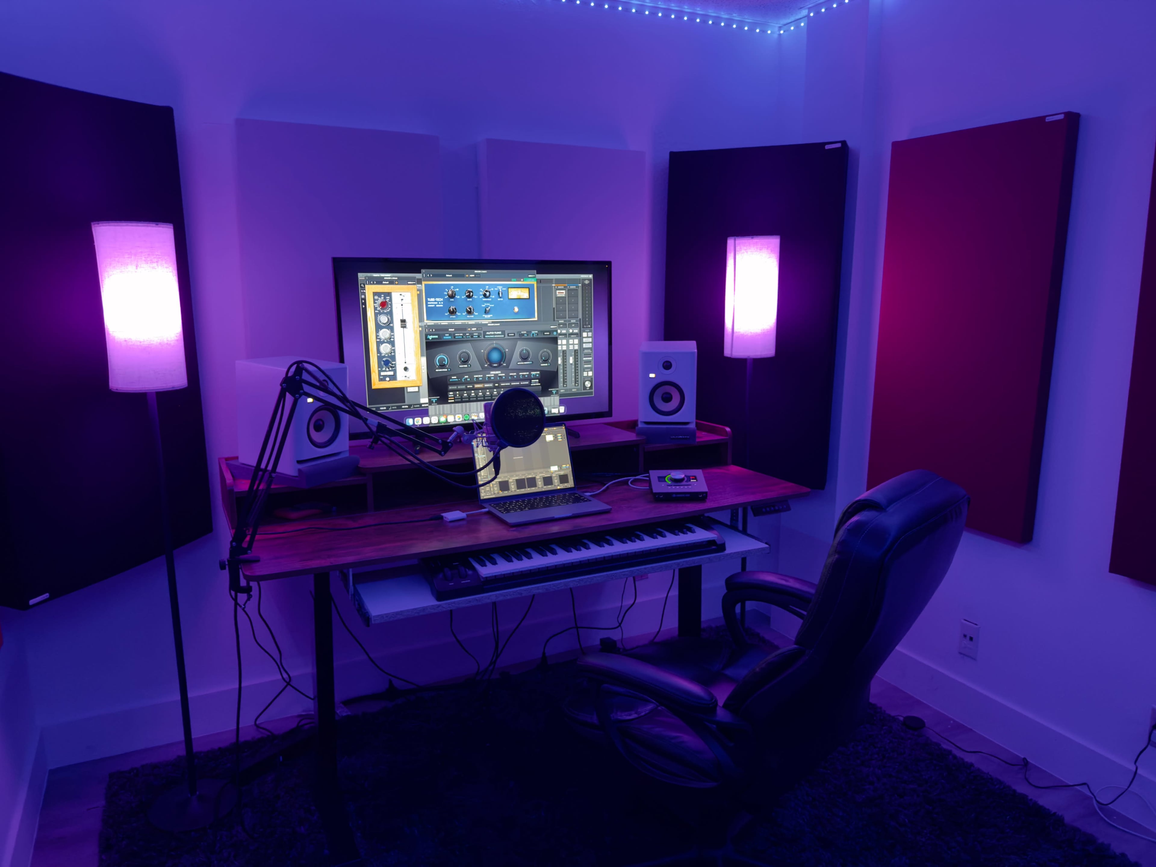 A home studio setup features a desk with a computer monitor, microphone, keyboard, and speakers, surrounded by acoustic panels and illuminated by purple lights.