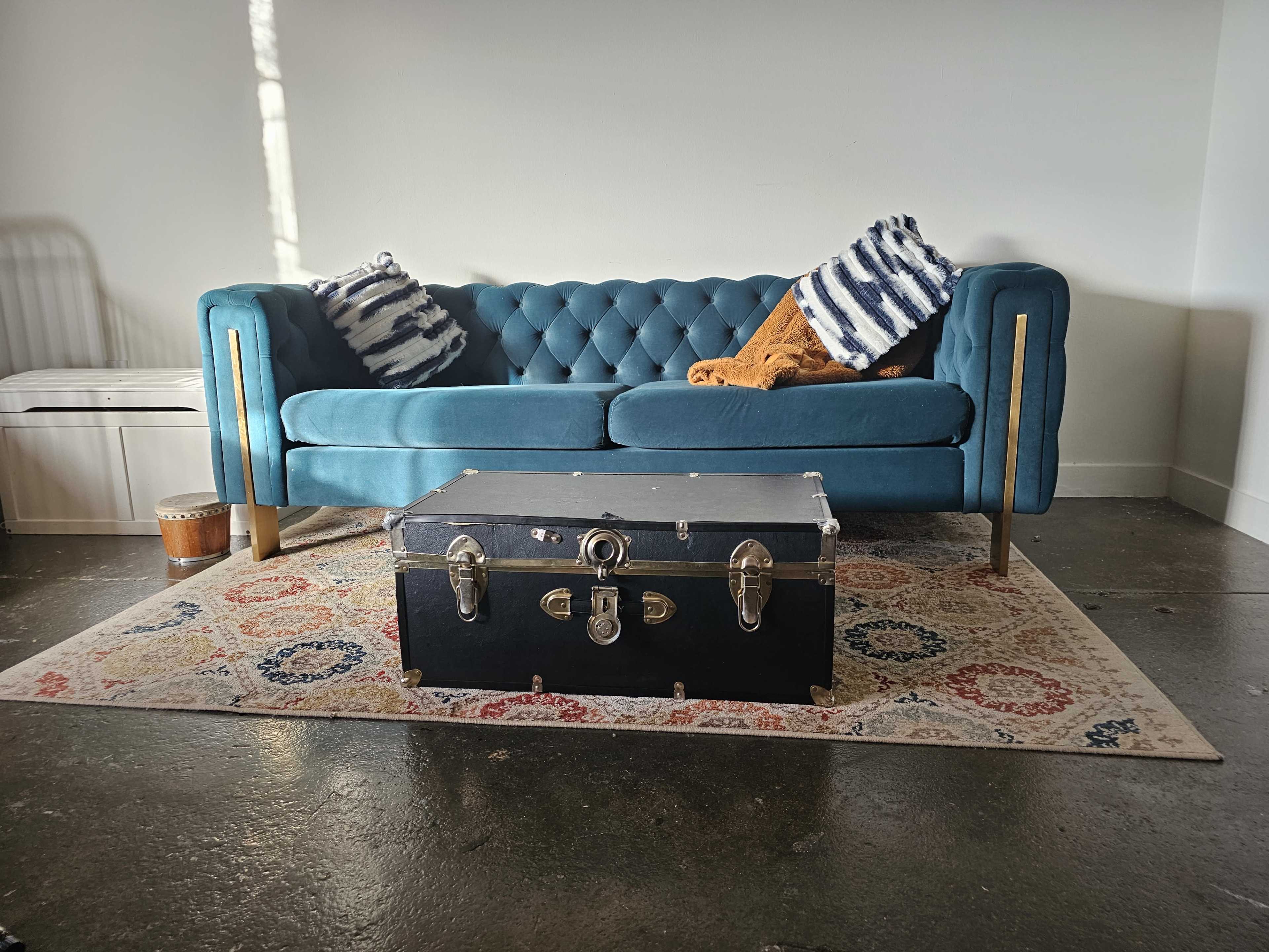 A tufted teal couch with striped pillows sits on a patterned rug, facing a vintage trunk in a bright room.