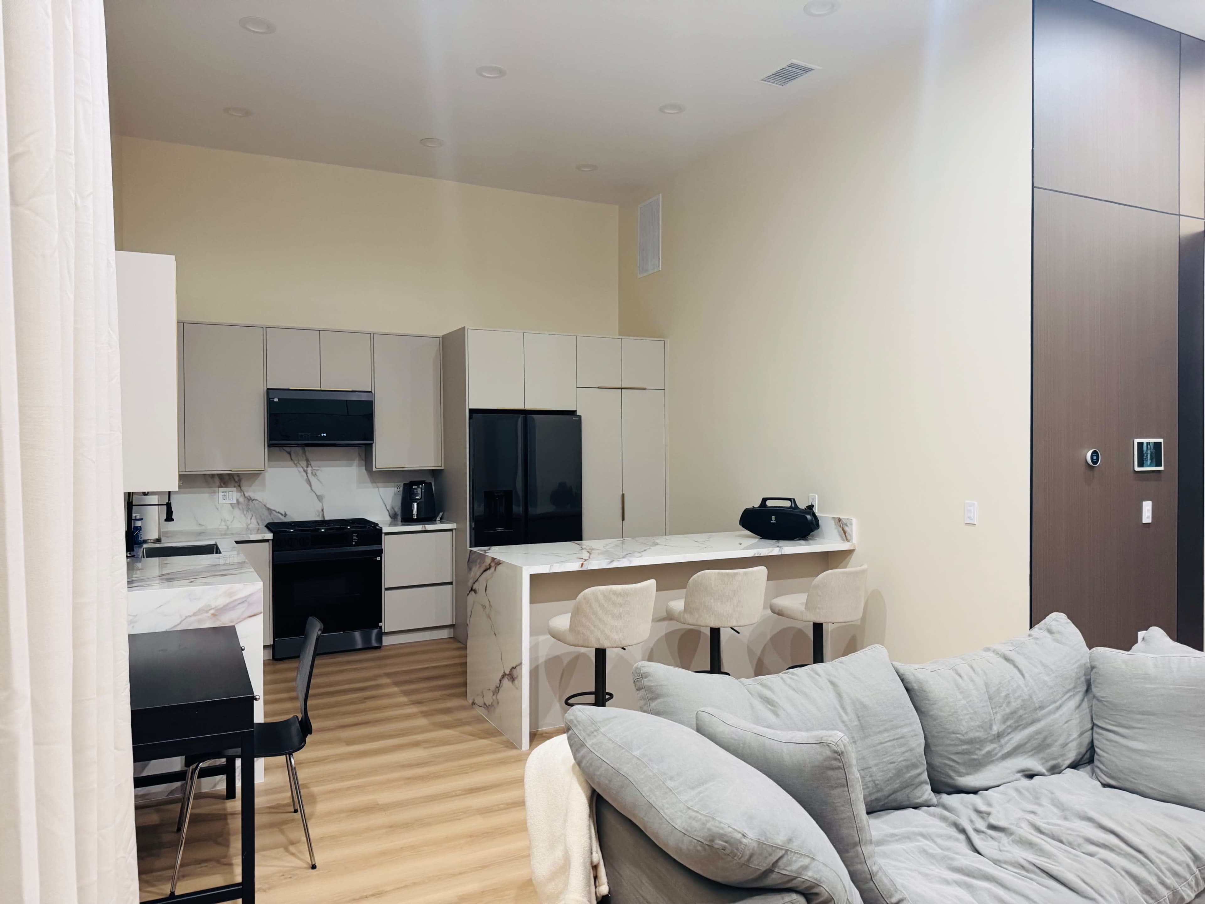 The image shows a modern kitchen and living area with a dining table, bar stools, and large windows.