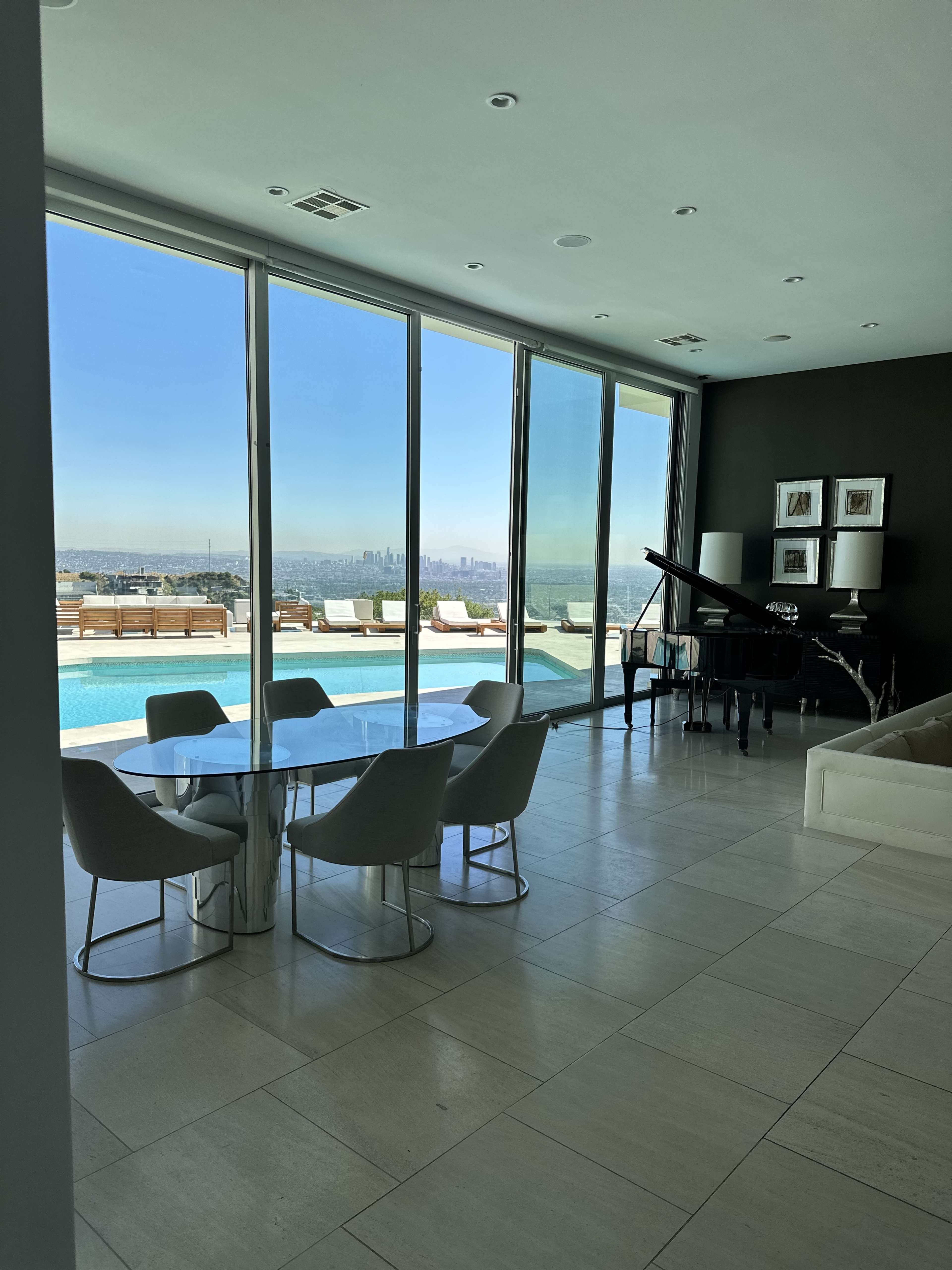 The image shows a modern interior with a glass dining table and chairs, a grand piano, and large windows overlooking a pool and city skyline.